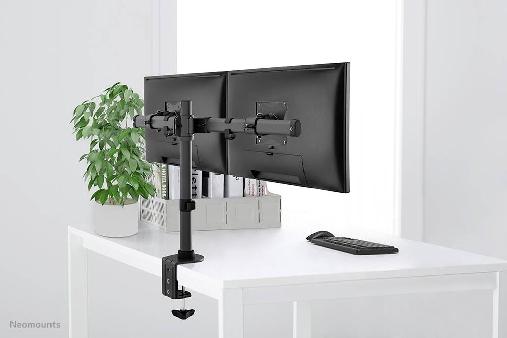 Ein Schreibtisch mit zwei nebeneinander montierten Monitoren, ein Tastaturset darunter, eine Pflanze links, in einem hellen Raum.