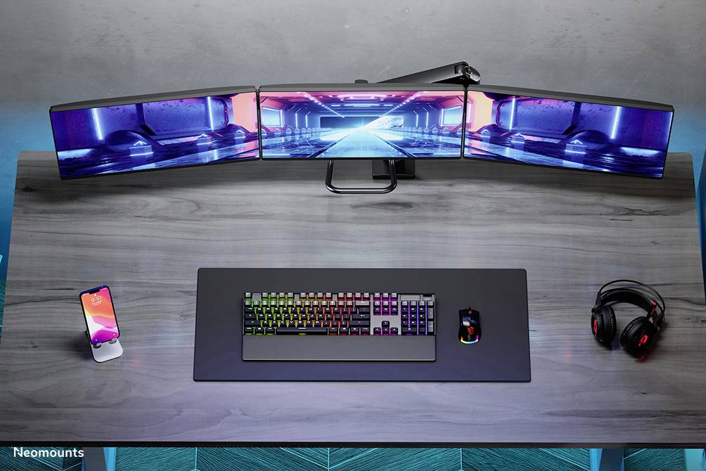 Ein Schreibtisch mit einer dreifachen Bildschirm-Setup, beleuchteter Tastatur, Maus, Smartphone und Kopfhörer; ideal für Gaming.