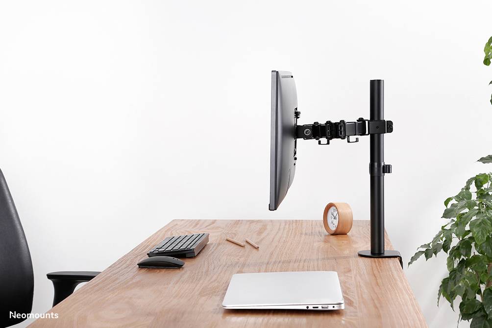 Monitorarm auf einem Schreibtisch, hält einen Bildschirm. Daneben Laptop, Tastatur, Maus, Uhr und Pflanzen im Hintergrund.