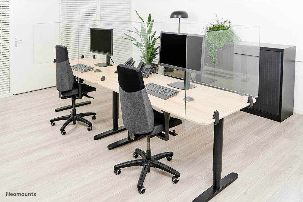 Zwei Büroarbeitsplätze mit Bildschirmen und Tastaturen, getrennt durch Plexiglas-Trennwände. Pflanzen stehen zwischen den Monitoren.