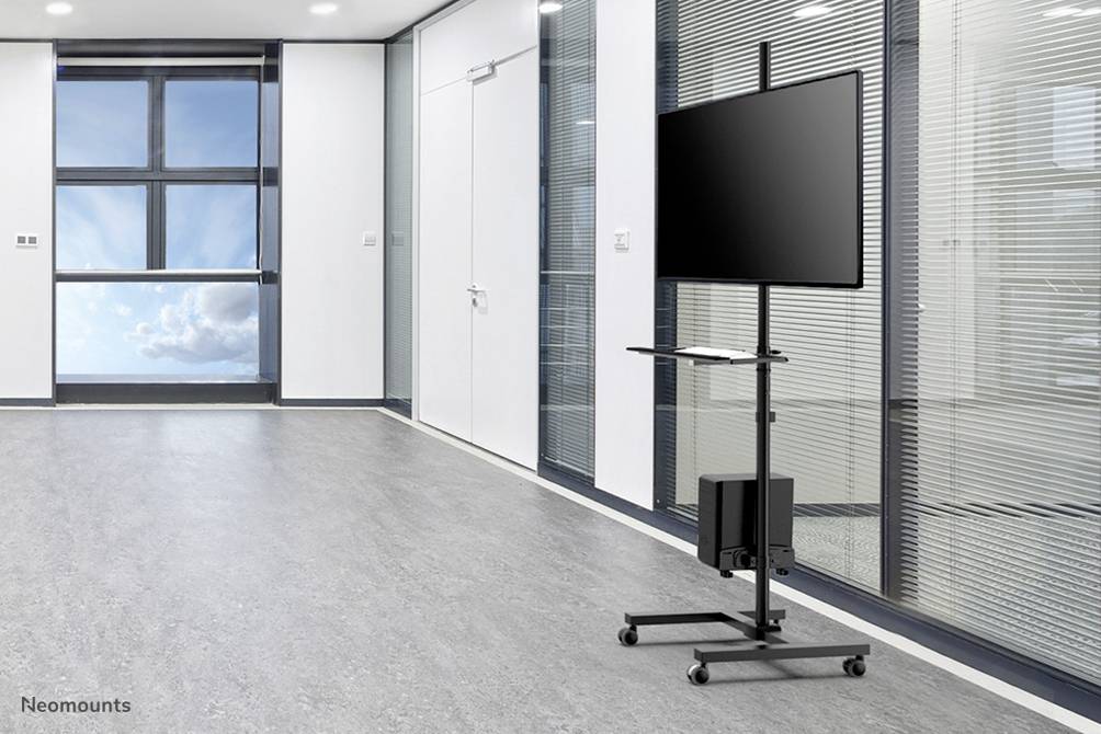 Ein leerer Konferenzraum mit großem Fenster und grauem Boden. Im Vordergrund steht ein mobiler Monitorständer mit einem Bildschirm.