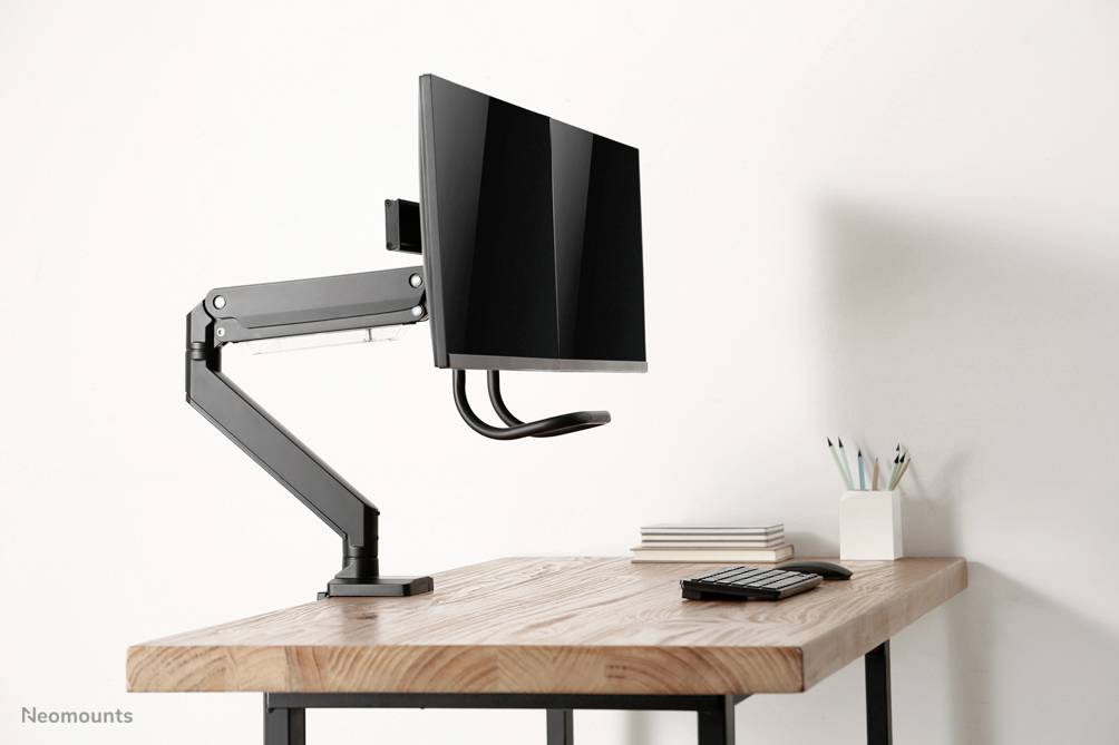 Ein Schreibtisch mit einem montierten Monitorarm, an dem ein Computerbildschirm hängt; daneben sind Stifte und Notizblöcke.