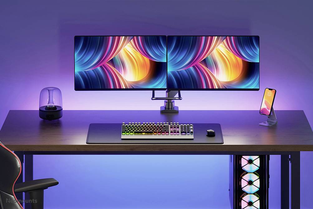 Ein Schreibtisch mit zwei leuchtend bunten Monitoren, einer RGB-Tastatur, einem Smartphone und einer Lampe, umgeben von blauer Hintergrundbeleuchtung.