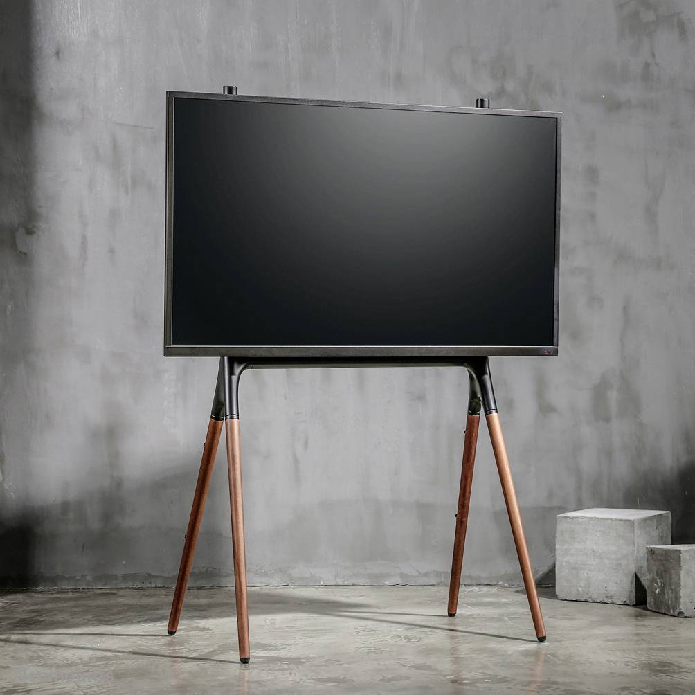 Ein moderner Fernseher auf einem dreibeinigen Holzständer steht in einem minimalistischen Raum mit Betonwänden.