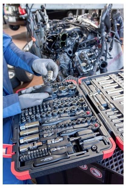 Die behandschuhten Hände eines Mechanikers sortieren Stecknüsse und Schraubenschlüssel neben einem zerlegten Automotor in einer Werkstatt.