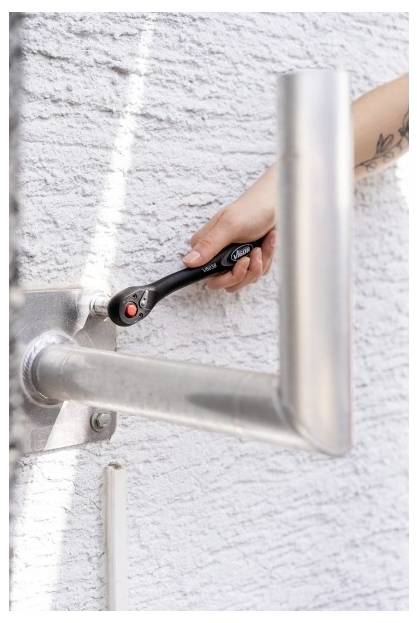 Das Handfestziehen eines schwarzen Ratschenwerkzeugs an einer an der Wand montierten Metallhalterung gegen eine weiße strukturierte Wand.