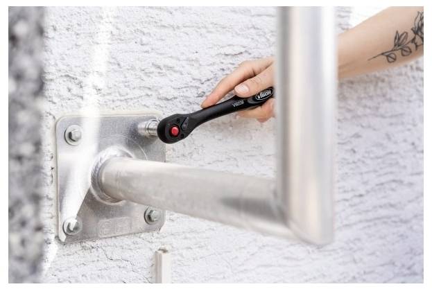 Eine Hand benutzt einen Schraubenschlüssel, um eine Metallhalterung festzuziehen, die ein Geländer an einer strukturierten Wand sichert.