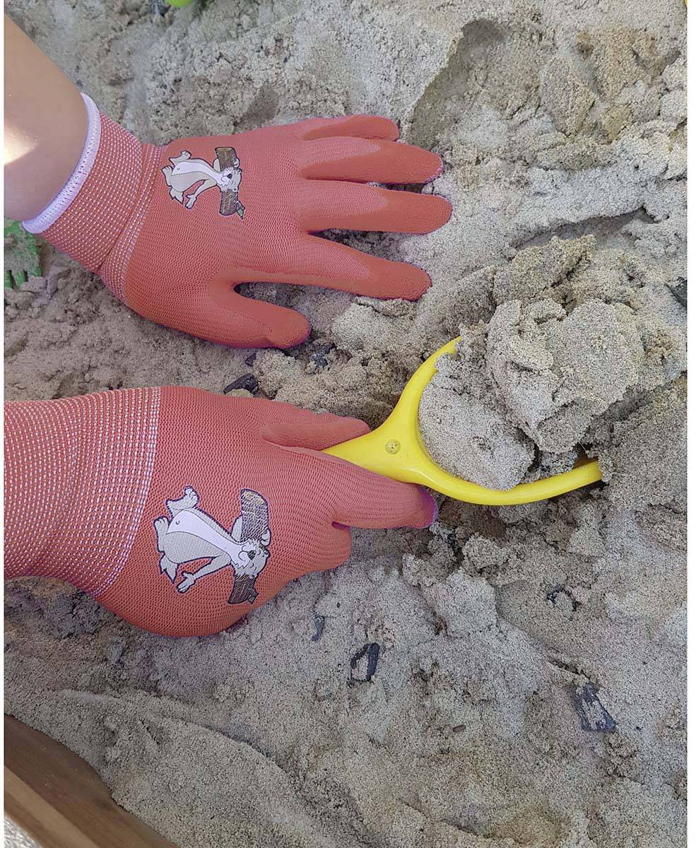 Ein Kind gräbt mit einer gelben Schaufel im Sand, trägt rote Handschuhe mit Hasenmotiv.