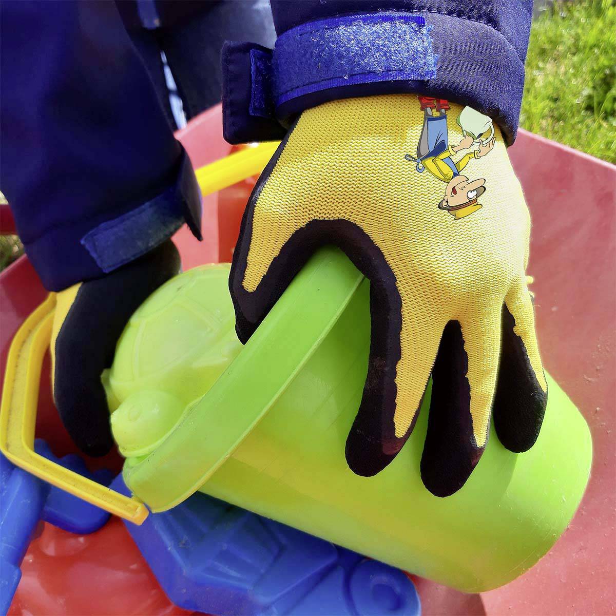 Ein bunte Handschuh hält einen grünen Eimer, umgeben von farbigem Sandspielzeug, auf einer Wiese.