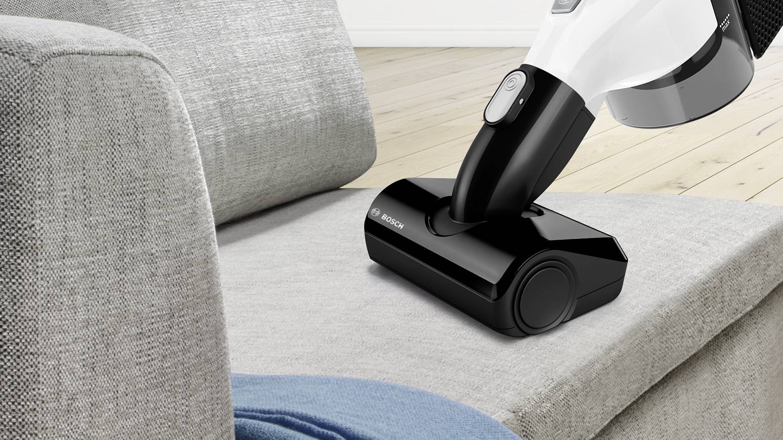 Schnurloser Staubsauger reinigt eine graue Sofaoberfläche im Wohnzimmer. Holzfußboden im Hintergrund.