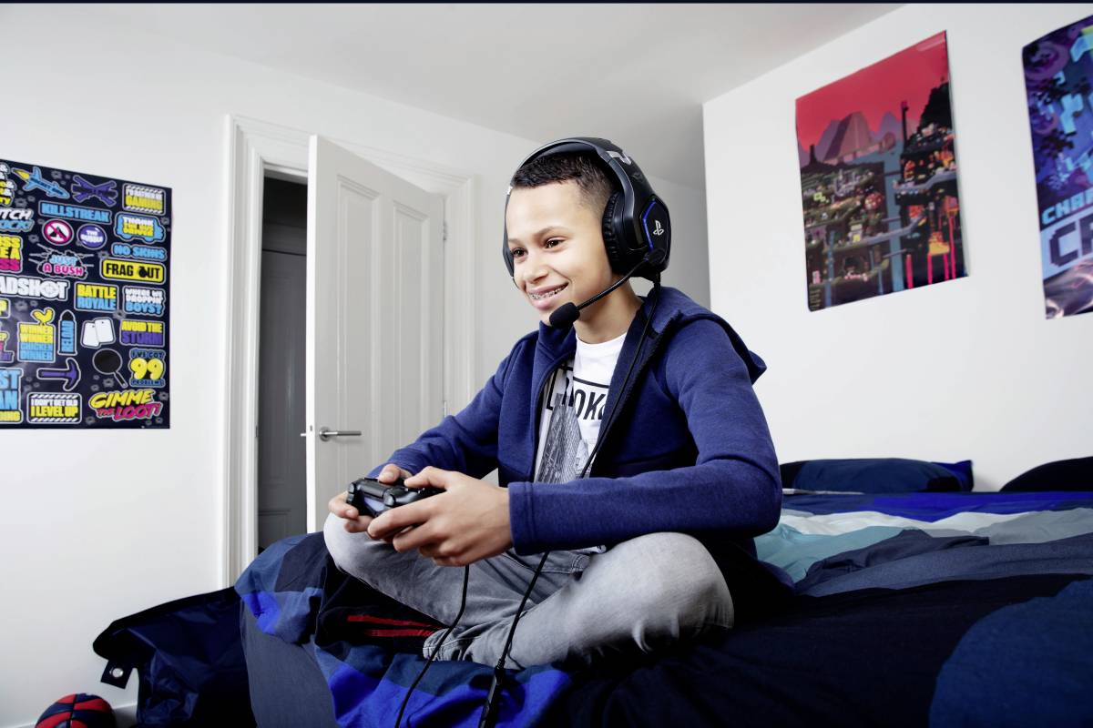 Junge spielt Videospiel im Schlafzimmer, trägt Headset, sitzt auf Bett, helle Wände mit Postern im Hintergrund.