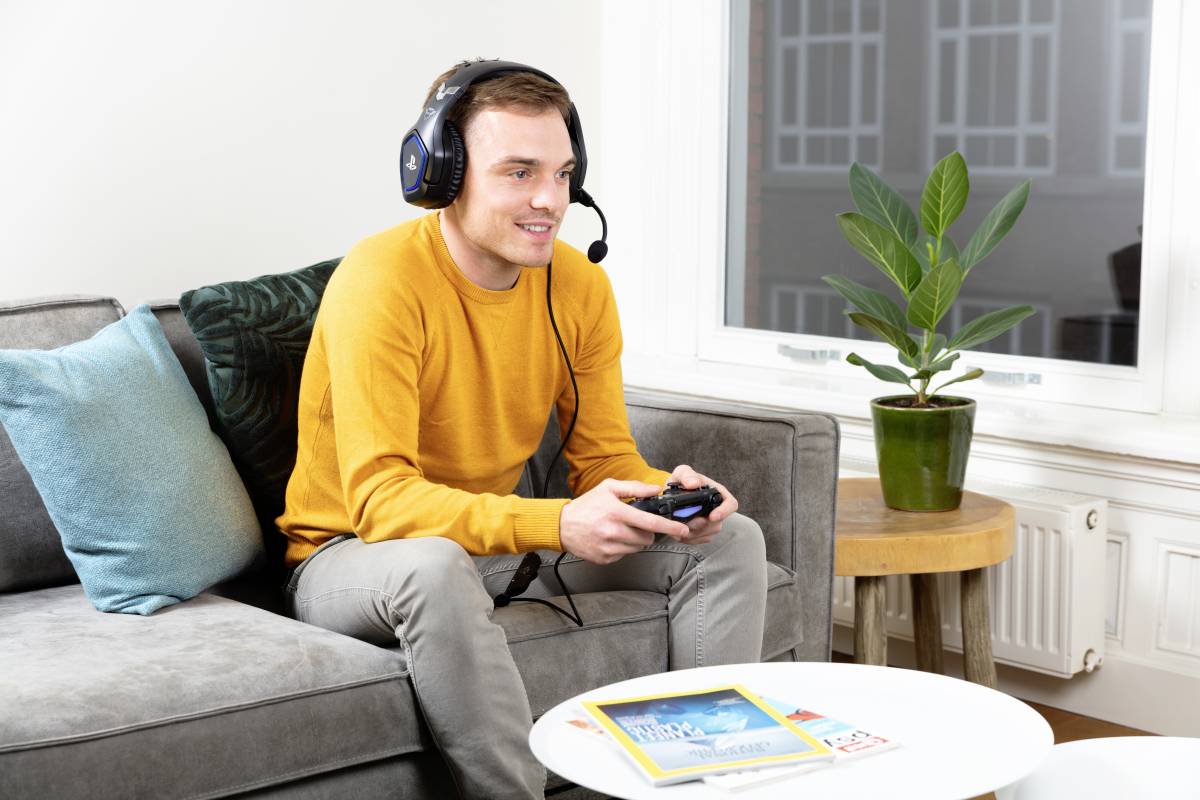 Ein Mann sitzt auf einem Sofa und spielt mit einem Controller, während er ein Headset trägt. Neben ihm liegt eine Zeitschrift auf dem Tisch.