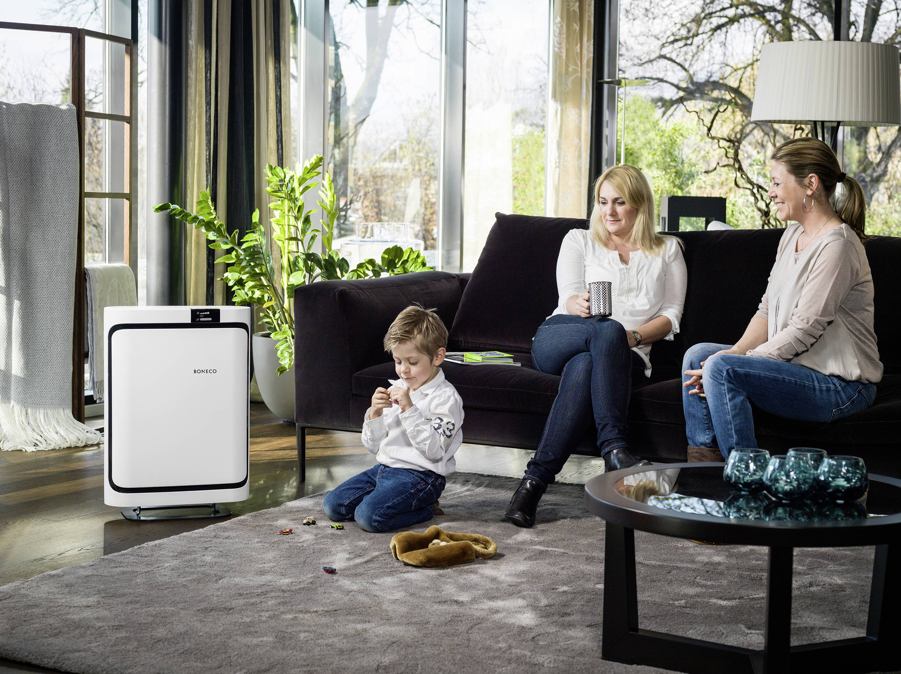 Ein Kind spielt auf dem Boden, während zwei Erwachsene auf einem Sofa sitzen und Kaffee trinken. Neben ihnen steht ein Luftreiniger.