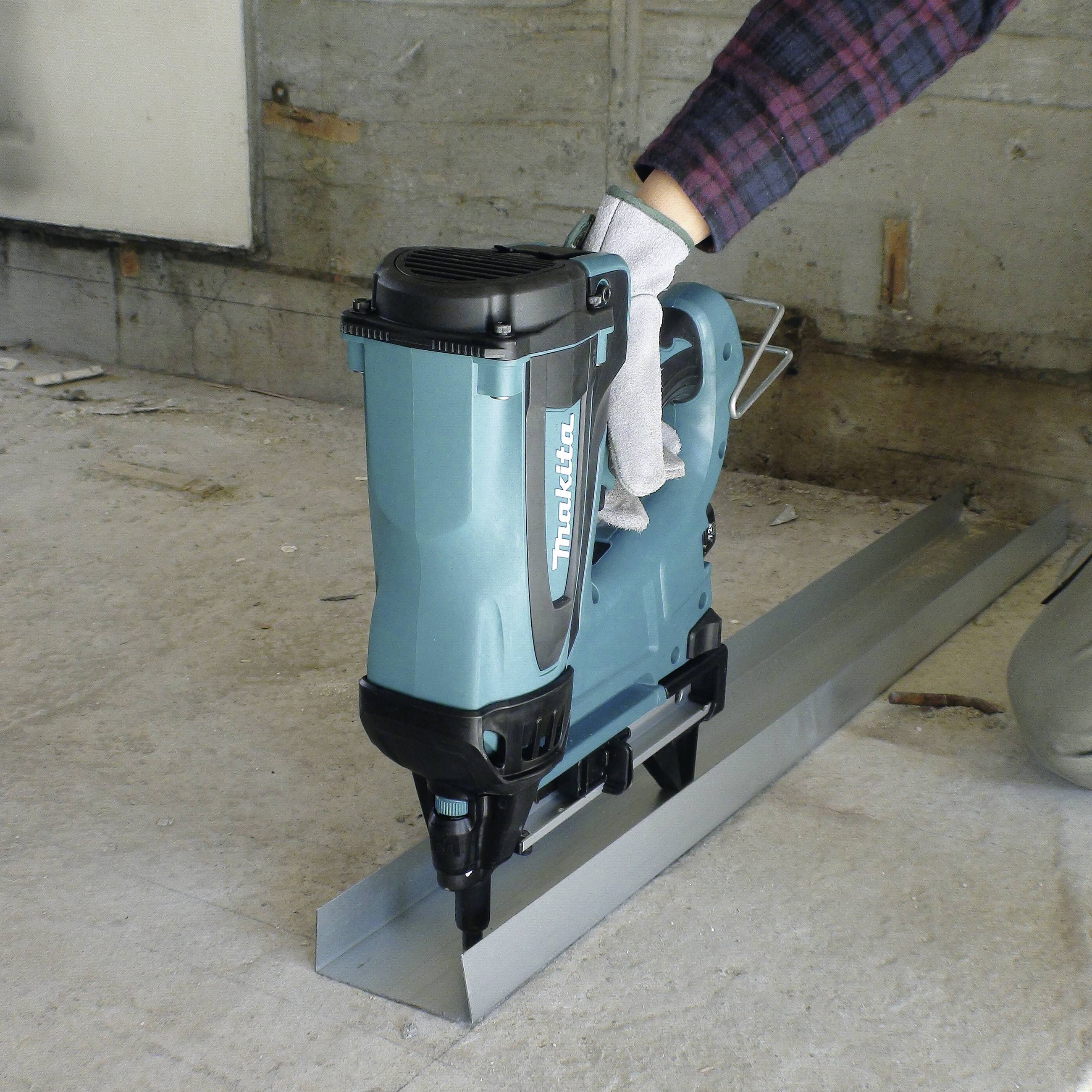 Eine Person hält ein türkisfarbenes Nagelgerät auf einem Metallstück auf einer Baustelle, sichtbar sind Betonwände im Hintergrund.