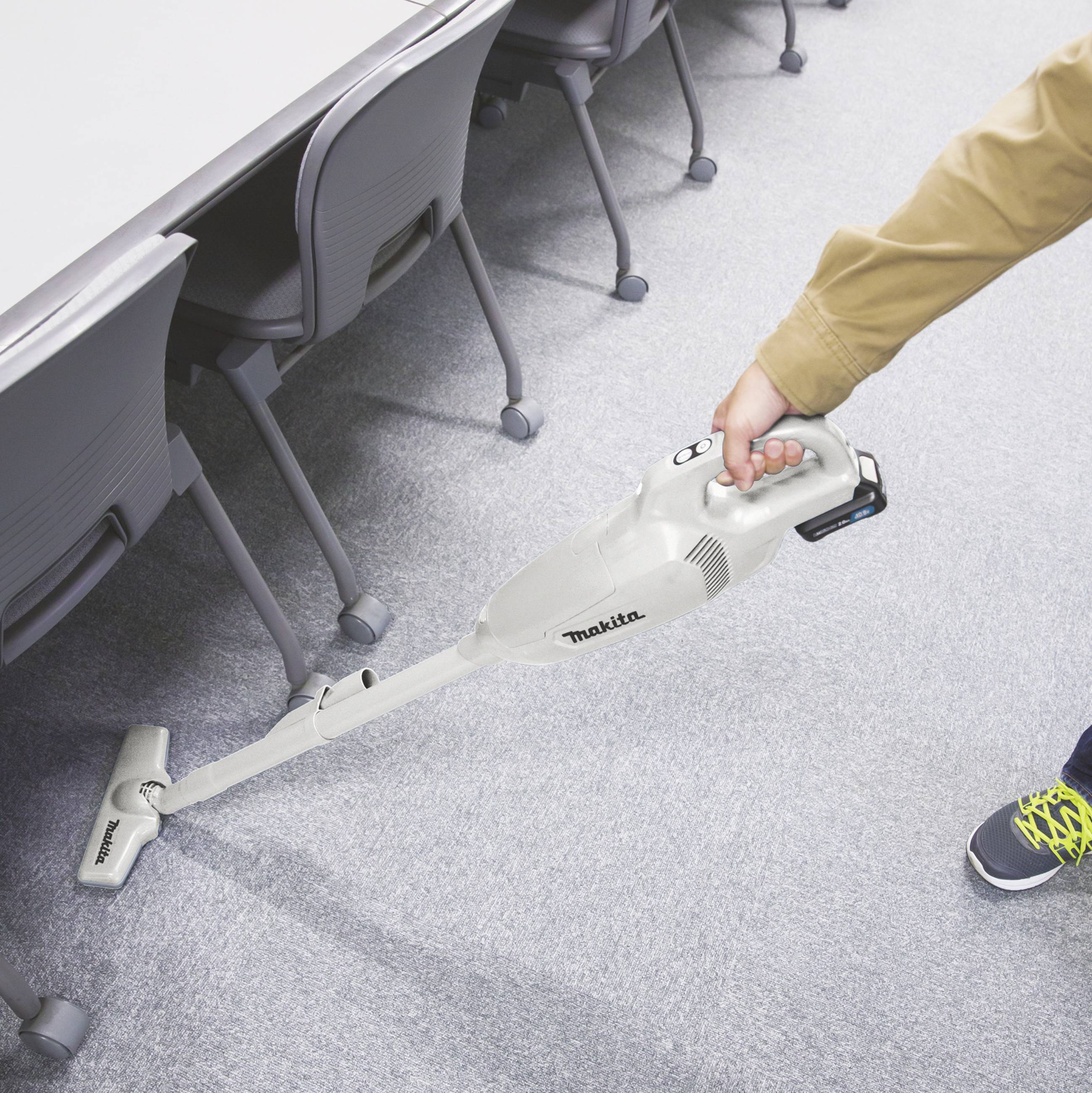 Person saugt einen Teppichboden neben einem Tisch mit einem kabellosen Staubsauger. Stuhlreihe in der Nähe, Enthüllung der Reinigungsaktivität.
