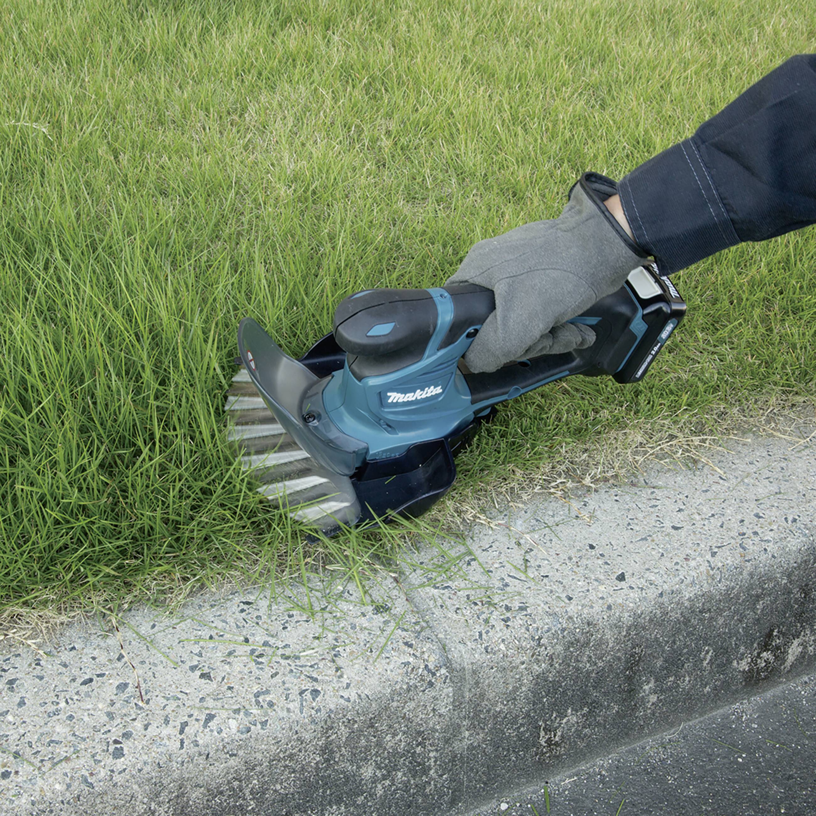 Eine Person schneidet mit einem kleinen, blauen elektrischen Rasentrimmer das Gras entlang eines Bordsteins.