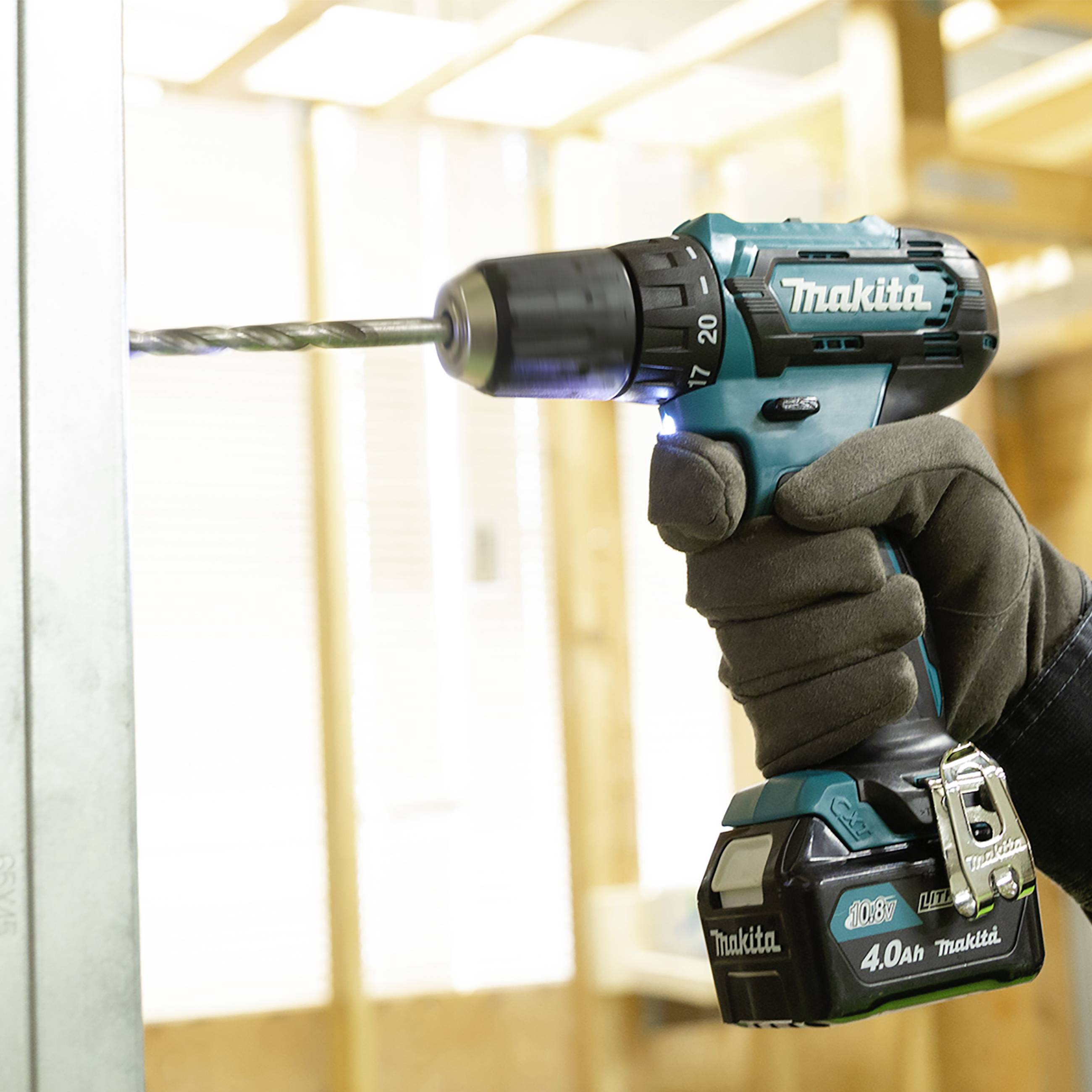 Eine Hand bohrt mit einer blauen Makita-Bohrmaschine in eine Holzwand. Im Hintergrund sind unscharfe Holzbalken zu sehen.
