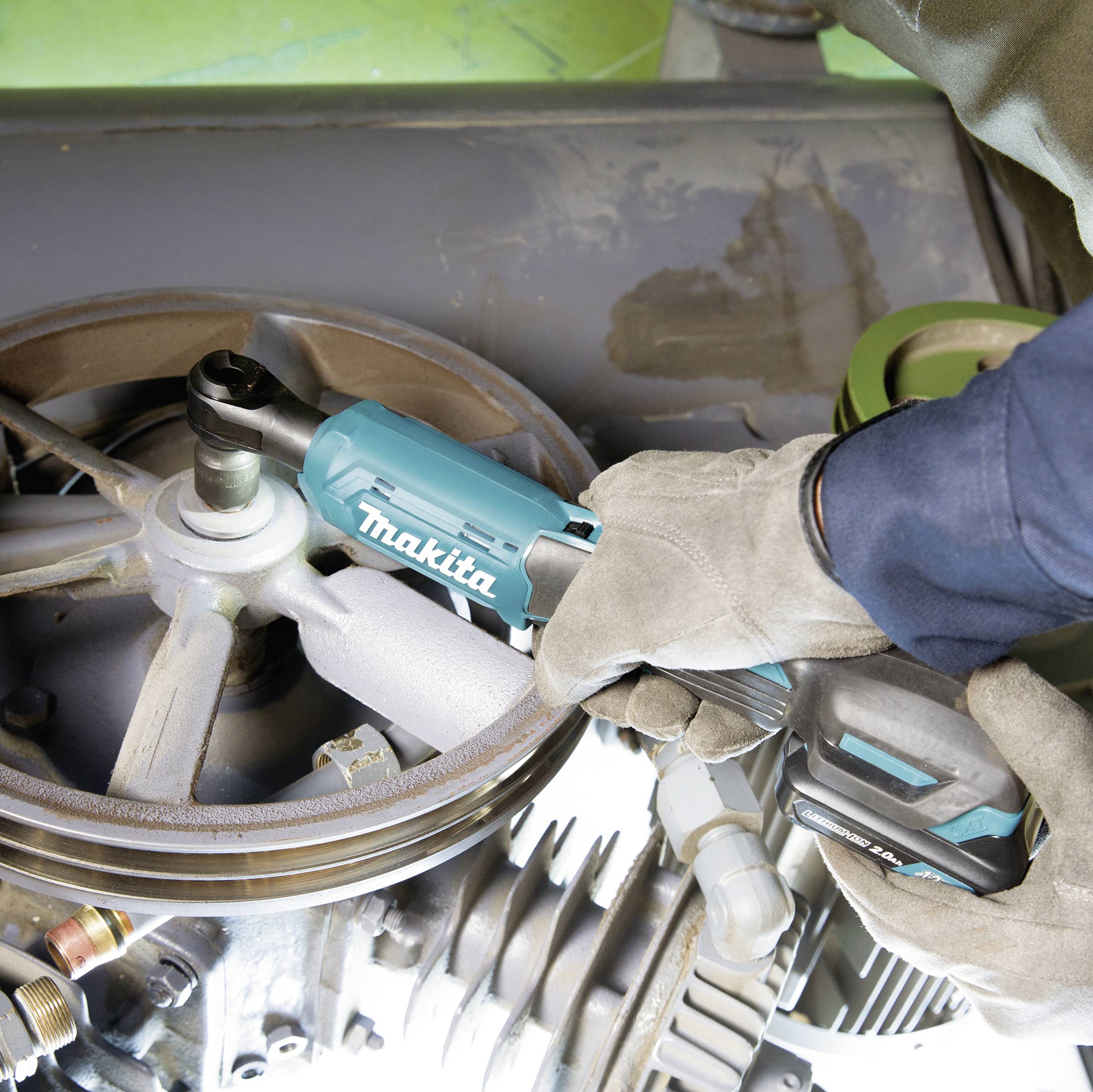 Ein Mechaniker mit Handschuhen verwendet ein elektrisches Werkzeug von Makita, um an einer großen Metallmaschine zu arbeiten.