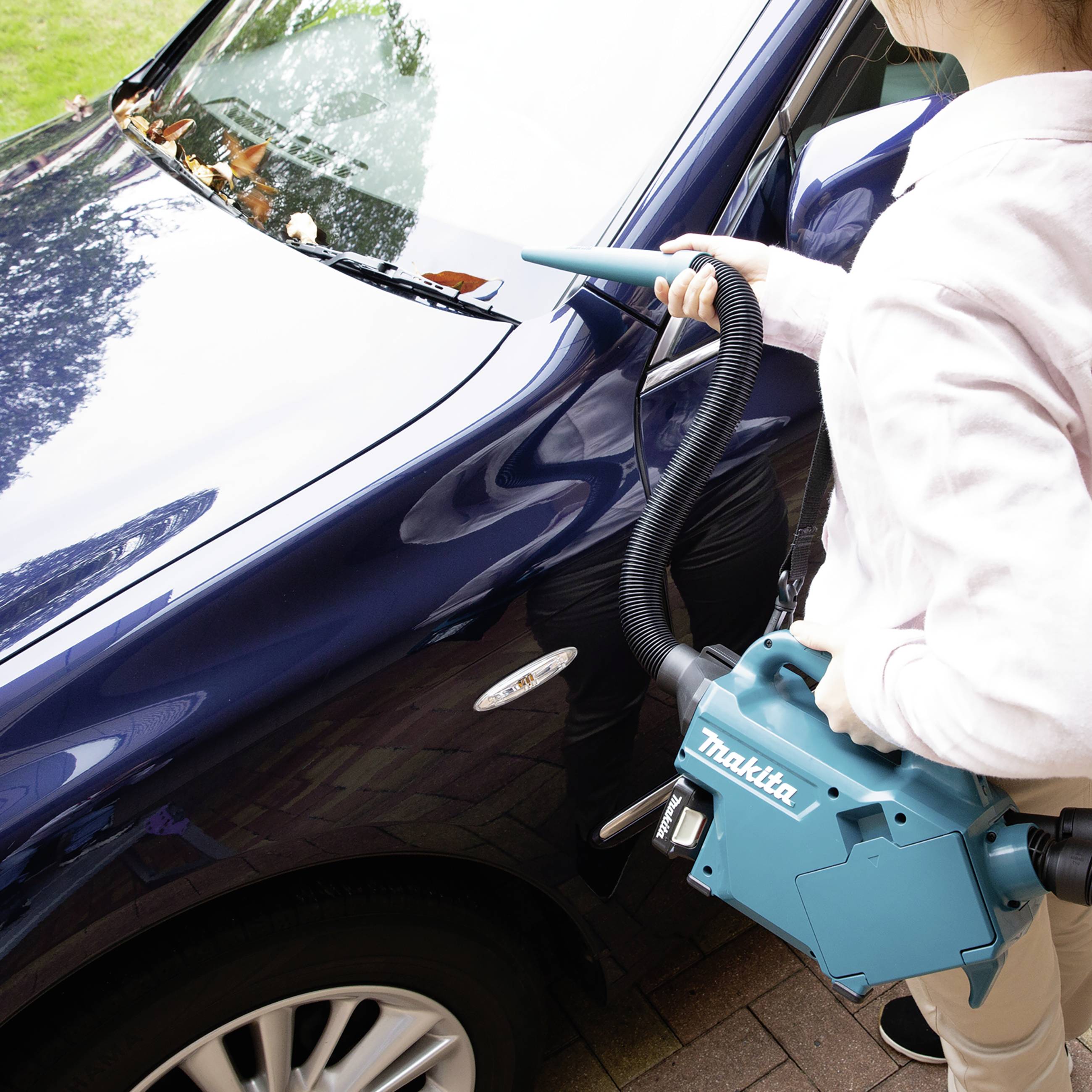 Person saugt Laub von der Windschutzscheibe eines blauen Autos mit einem tragbaren Staubsauger ab. Der Staubsauger ist türkisfarben.