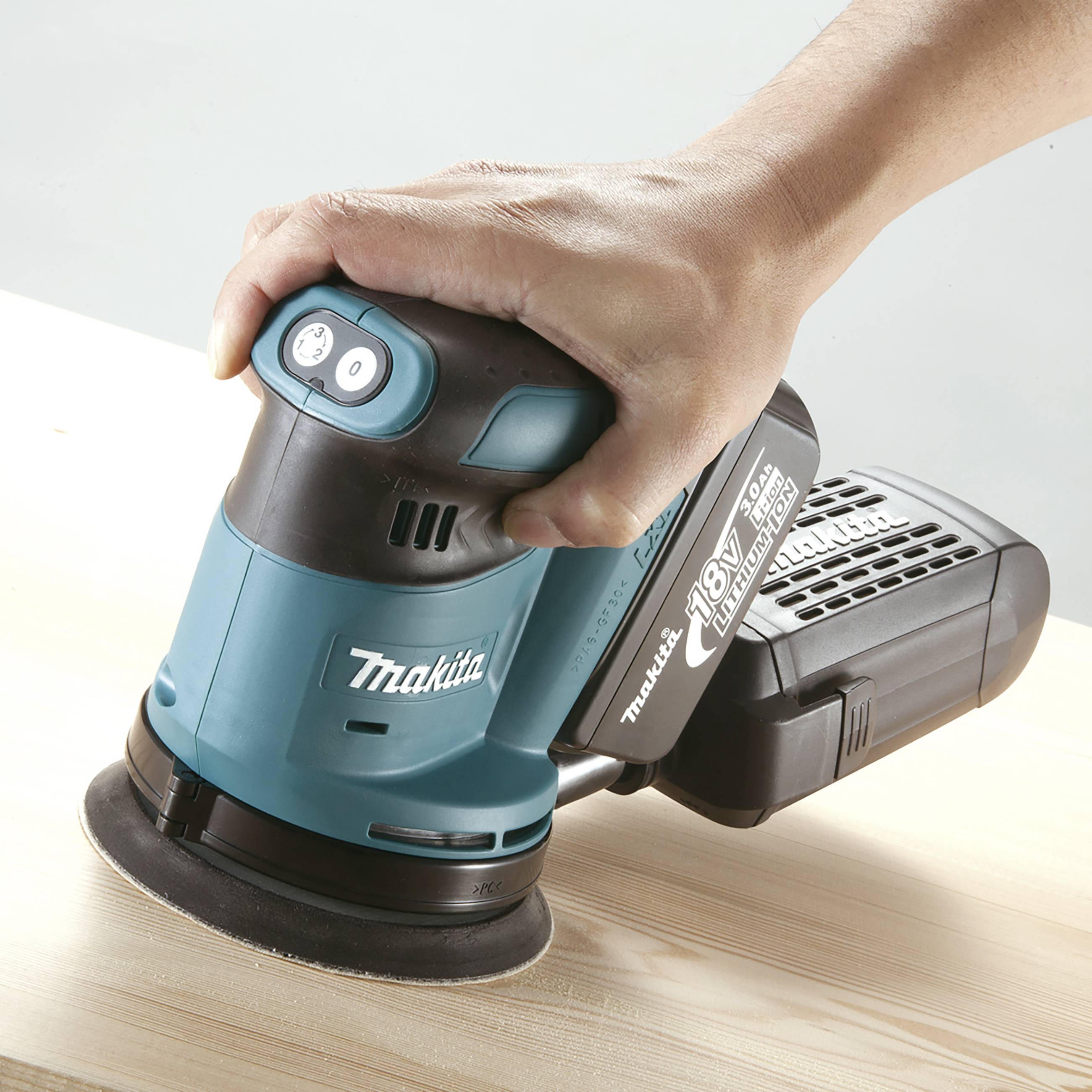 Eine Hand hält einen blauen Schleifer der Marke Makita, während er über eine Holzoberfläche betrieben wird.