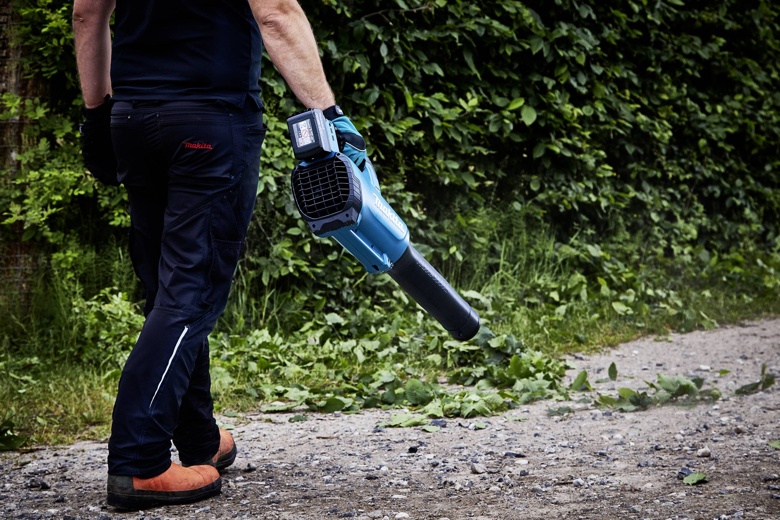 Person bläst Laub mit einem elektrischen Laubbläser von einem Schotterweg. Im Hintergrund ist grüne Vegetation zu sehen.