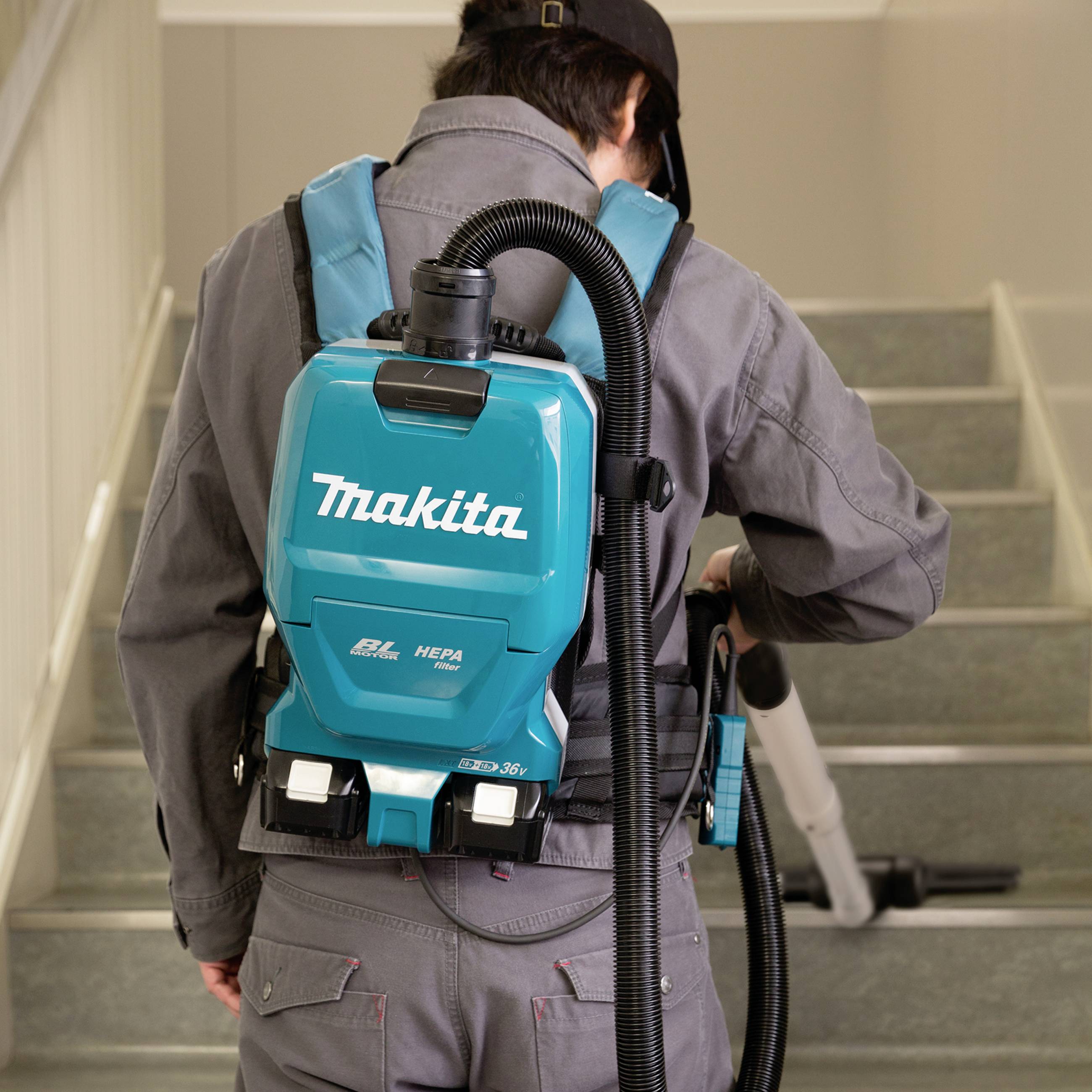 Eine Person reinigt eine Treppe mit einem Rucksackstaubsauger. Der Staubsauger ist hellblau und trägt das Logo 'Makita'.