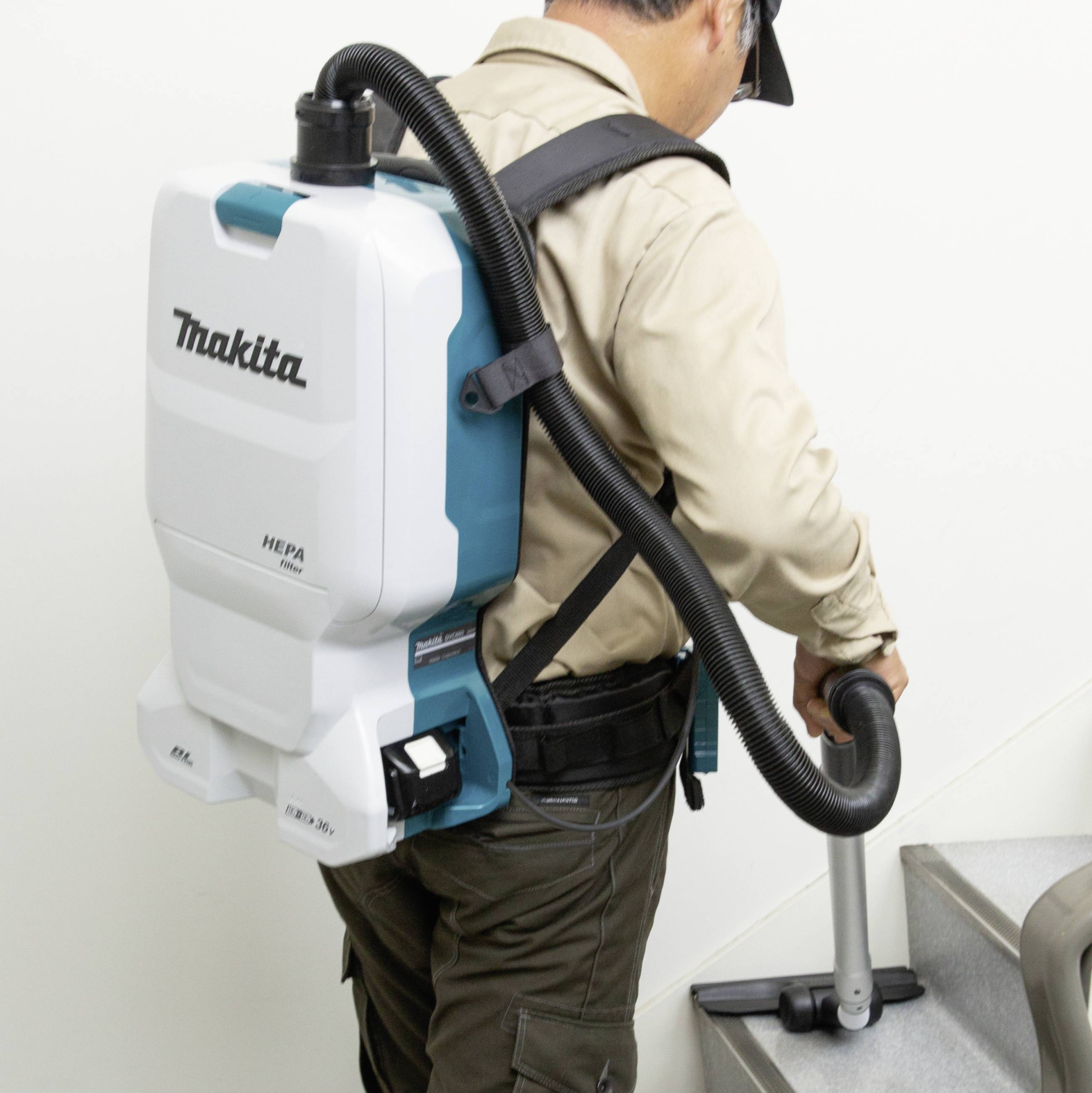 Eine Person verwendet einen tragbaren Staubsauger auf einer Treppe. Der Staubsauger ist auf dem Rücken befestigt und wird von der Marke Makita hergestellt.