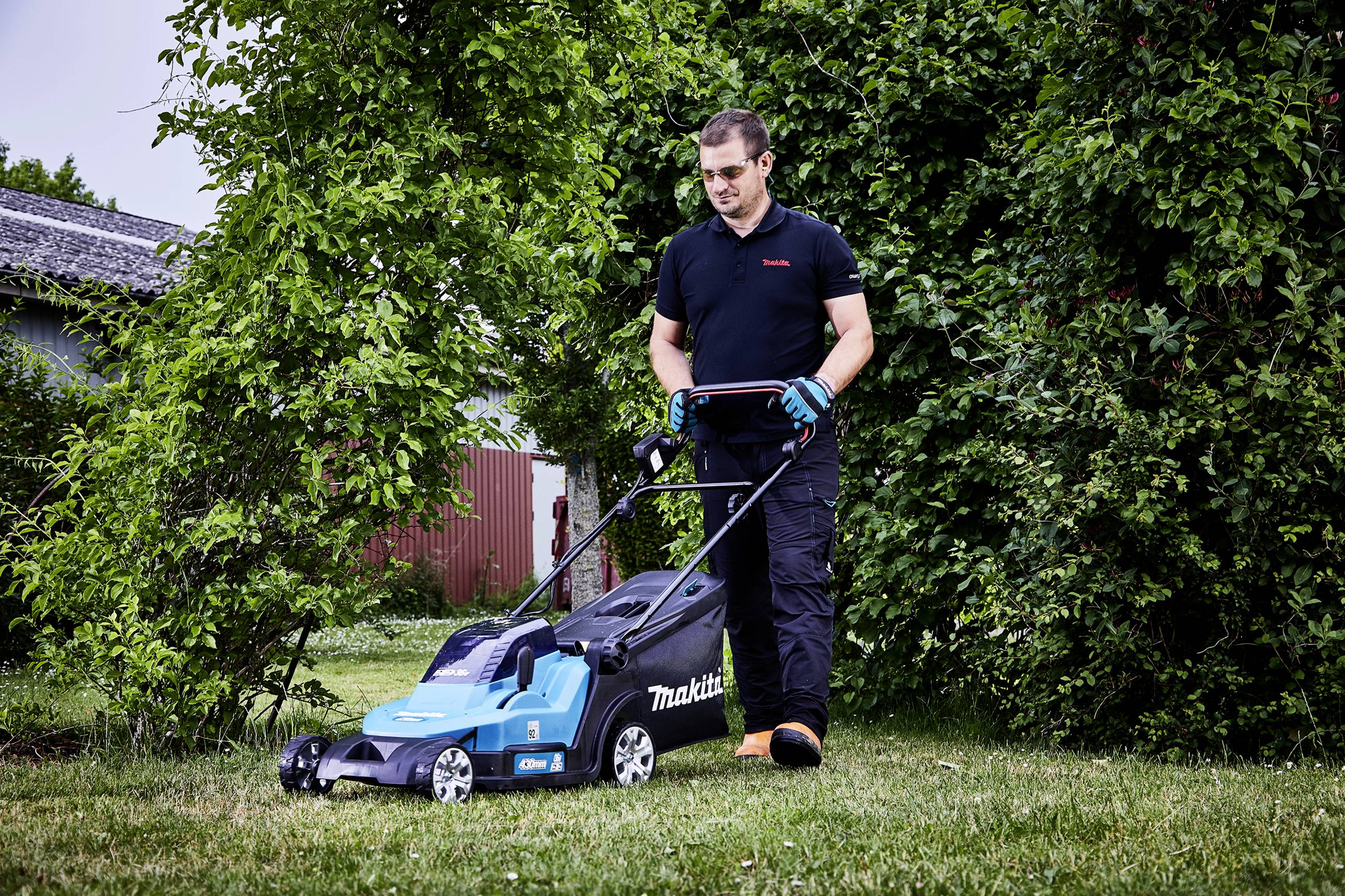 Ein Mann mäht einen Rasen in einem Garten mit einem blauen Rasenmäher. Im Hintergrund sind Büsche und ein Gebäude zu sehen.