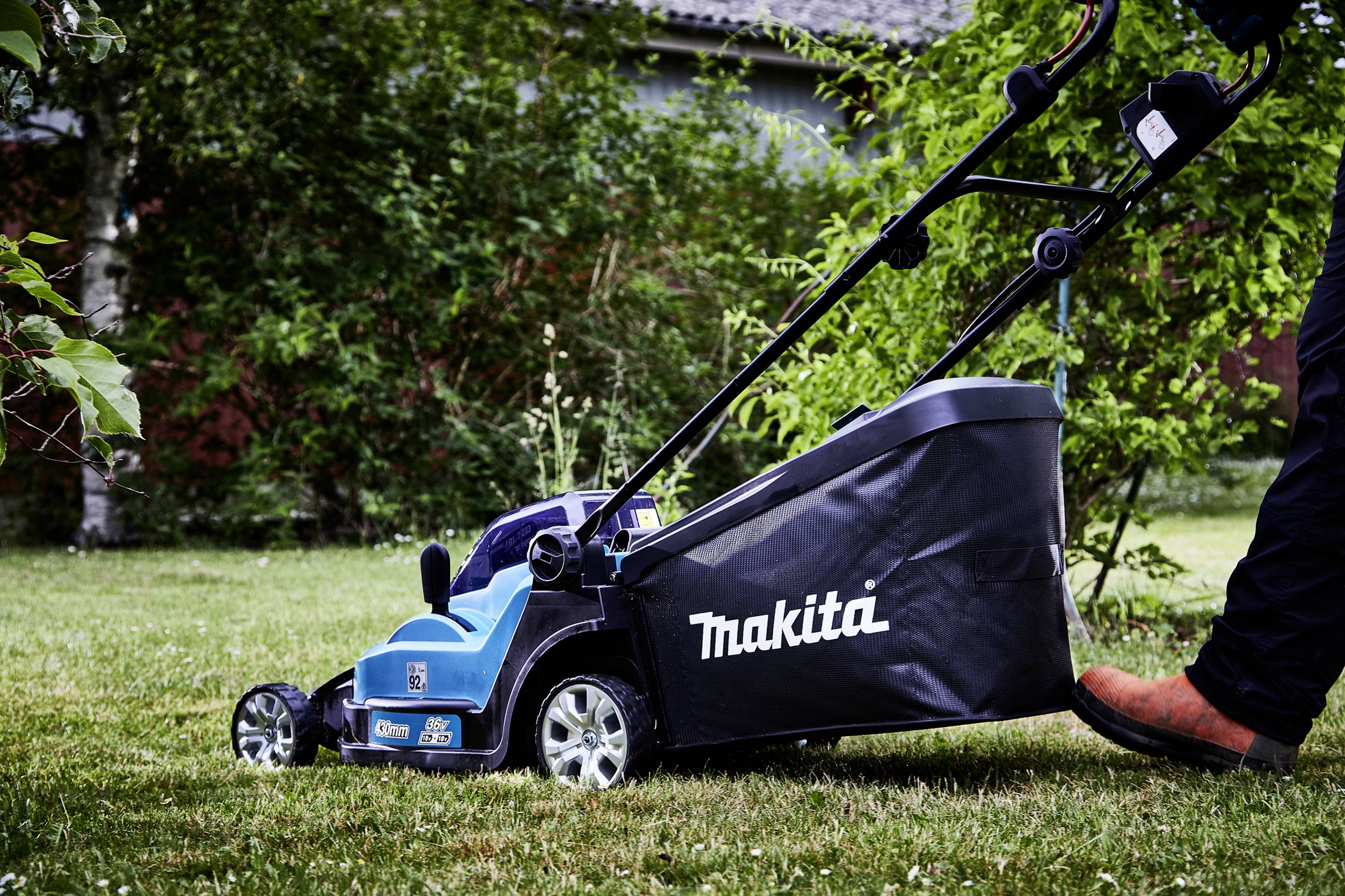Rasenmäher der Marke Makita wird auf einem grünen Rasen benutzt. Eine Person schiebt den Rasenmäher, während sie festes Schuhwerk trägt.