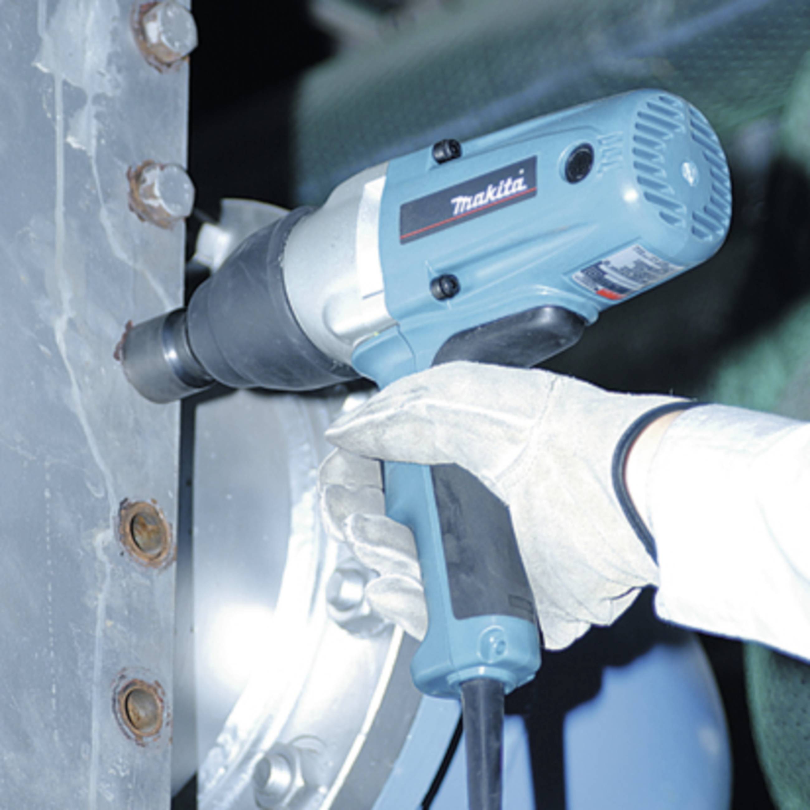 Eine Person in weißen Arbeitshandschuhen benutzt eine blaue elektrische Bohrmaschine. Die Szene zeigt industrielles Arbeiten an Metall.