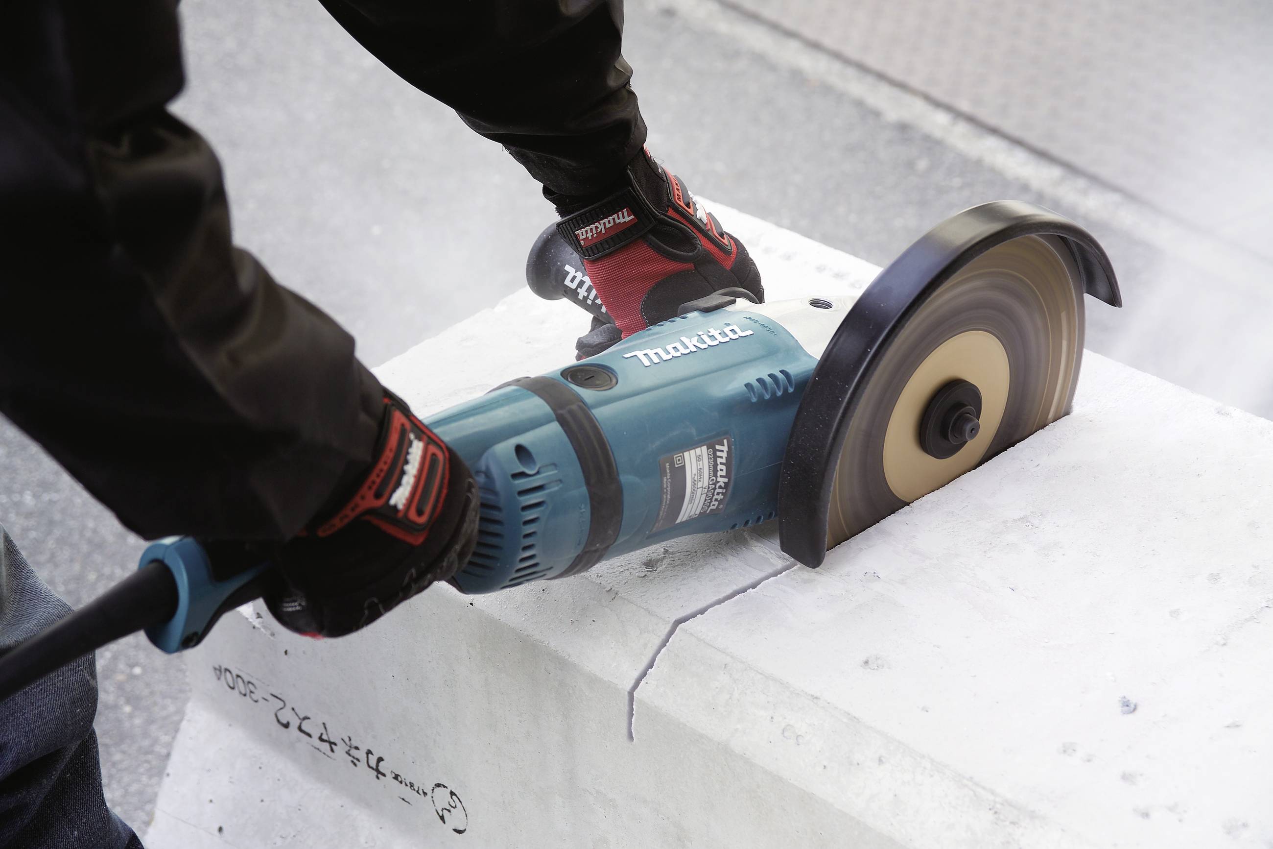 Eine Person schneidet mit einer Handkreissäge auf einem Betonblock, trägt Handschuhe und Schutzkleidung; Staub wird aufgewirbelt.