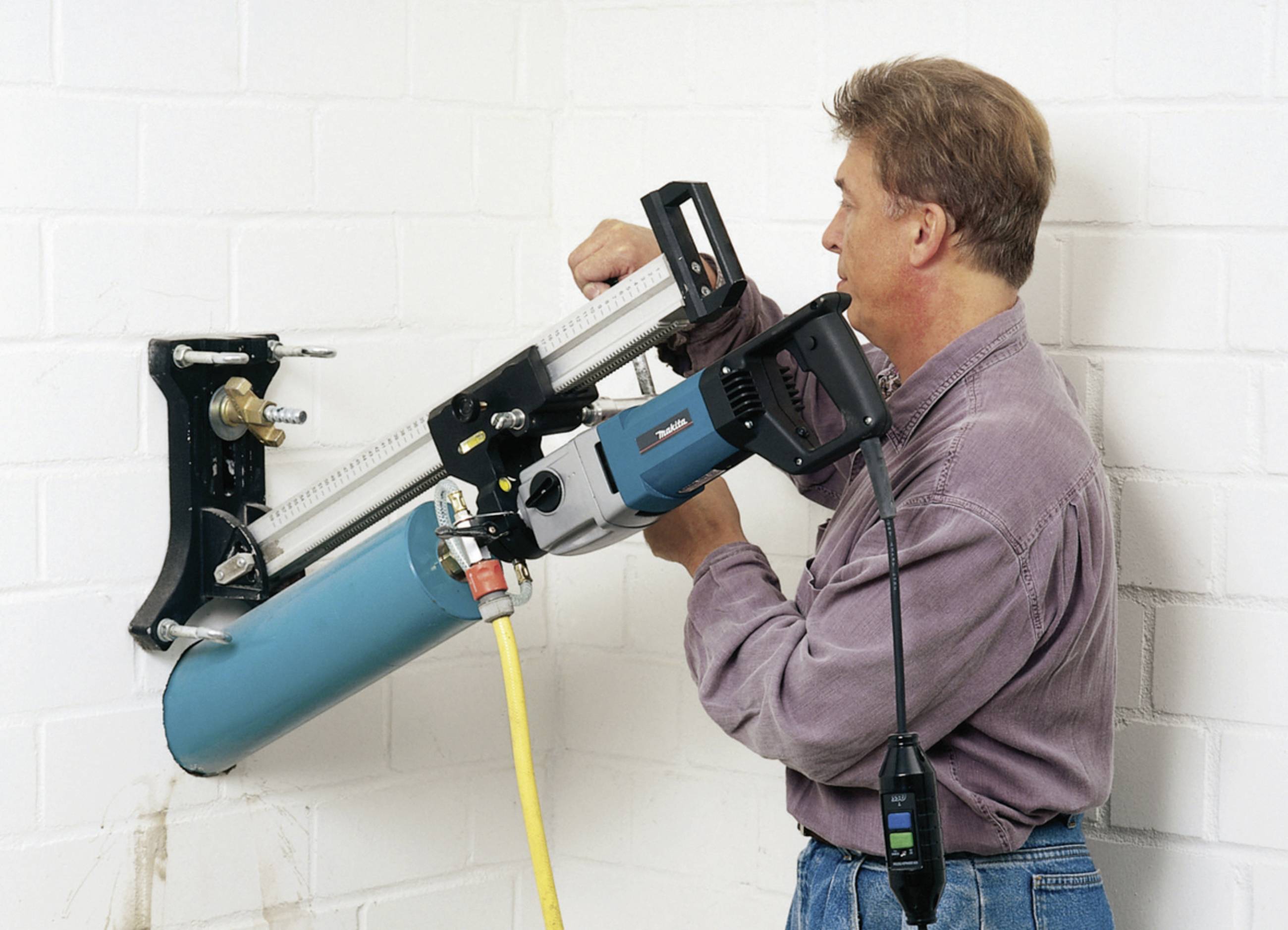 Ein Mann benutzt eine Bohrmaschine, um ein großes Loch in eine weiße Mauer zu bohren. Er trägt ein graues Hemd und konzentriert sich auf die Arbeit.