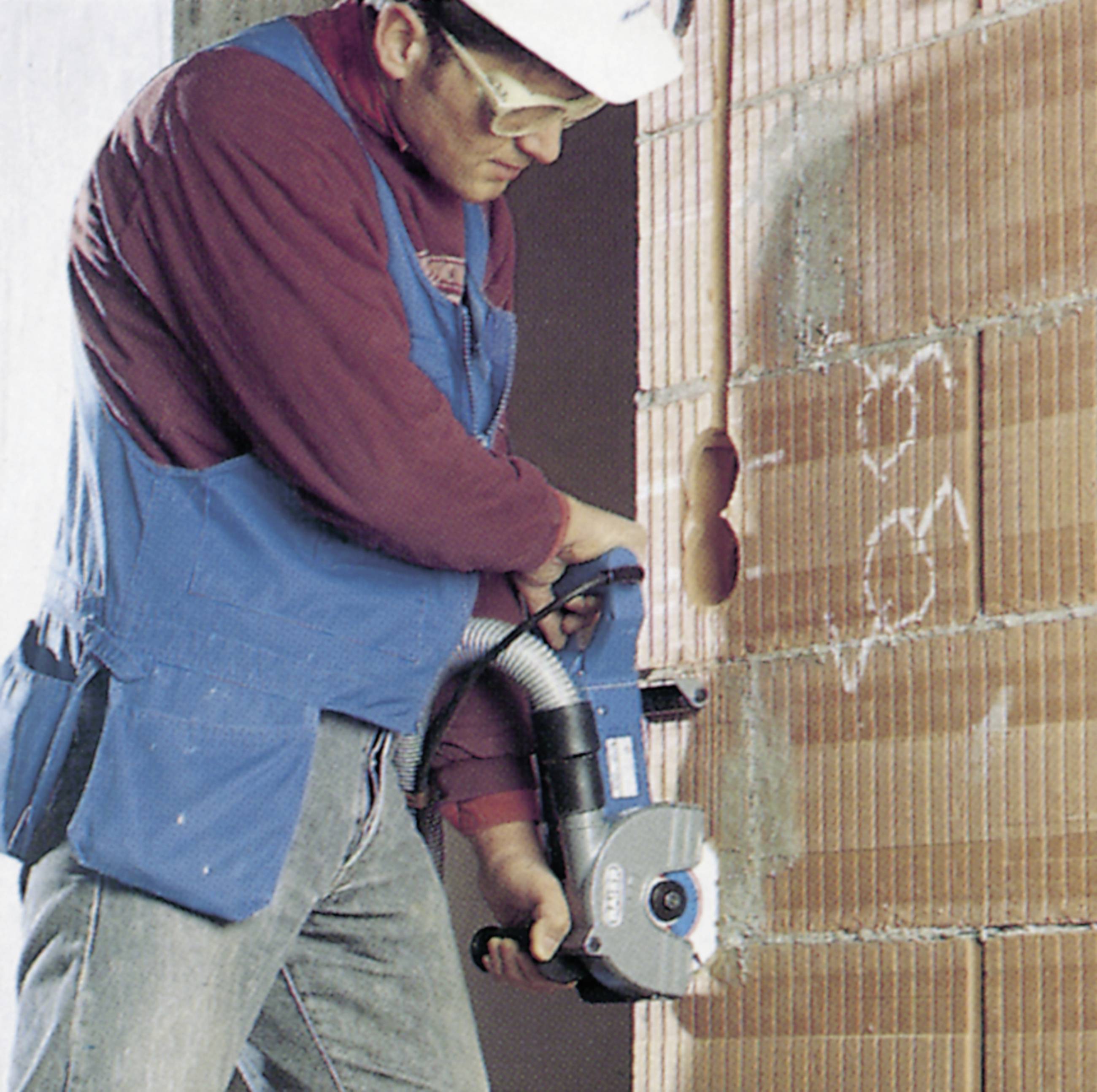 Ein Bauarbeiter in Schutzkleidung schneidet mit einer Säge Fugen in eine Ziegelwand.