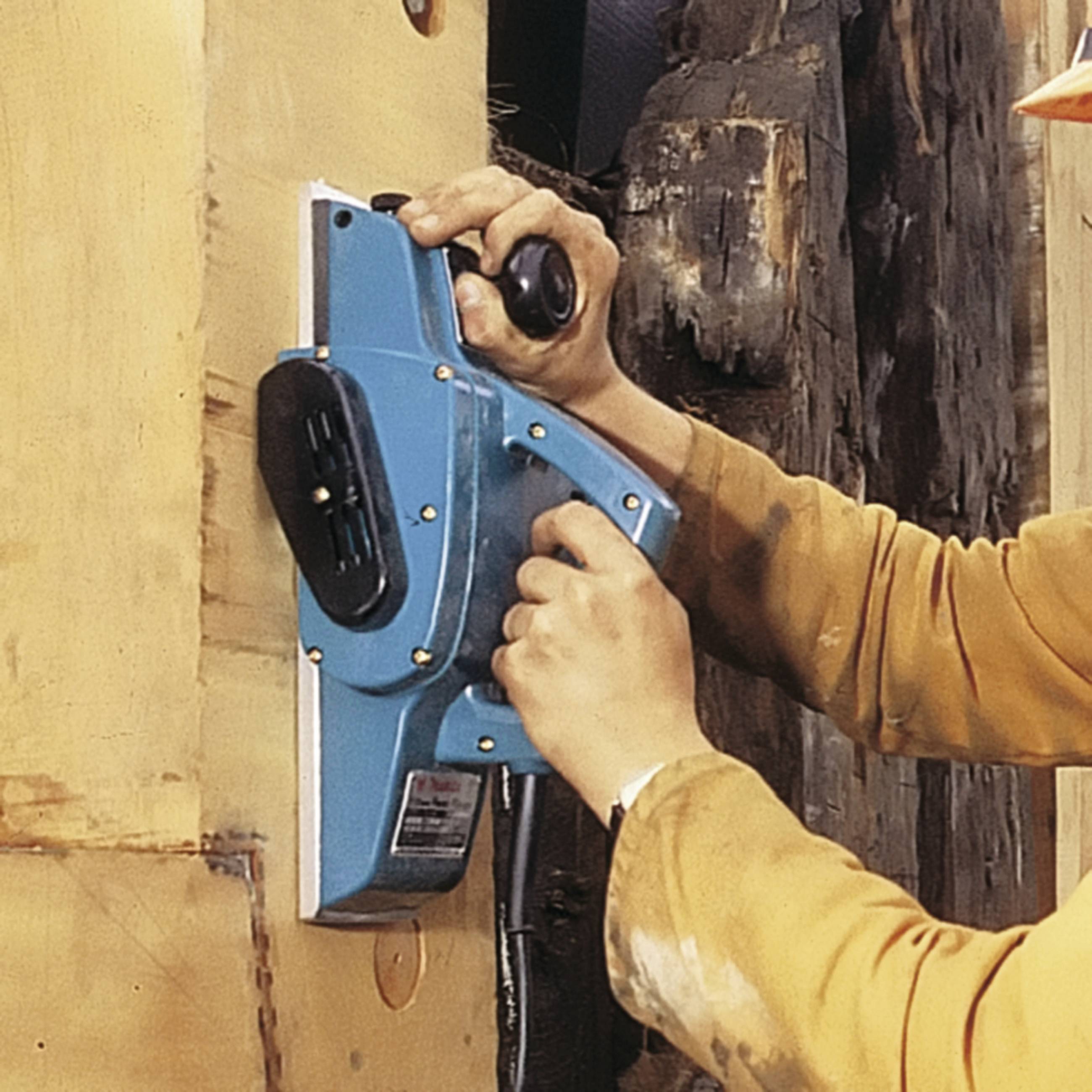 Person schleift Holz mit einer elektrischen Schleifmaschine. Grob strukturiertes Holz ist sichtbar. Arbeitsumgebung wirkt handwerklich.