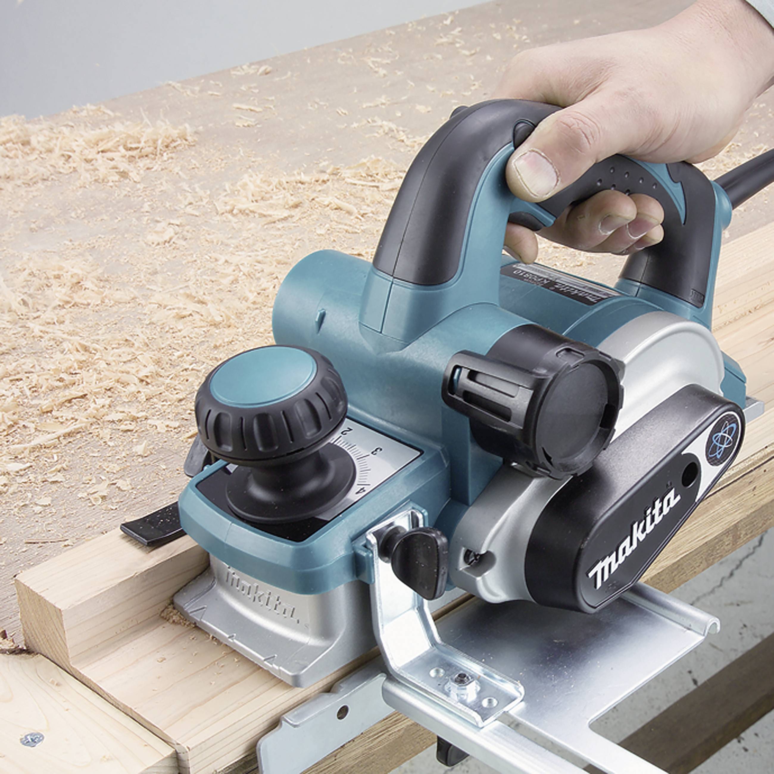 Eine Hand führt eine blaue Elektrohobelmaschine über eine Holzoberfläche, mit Holzspänen im Vordergrund.