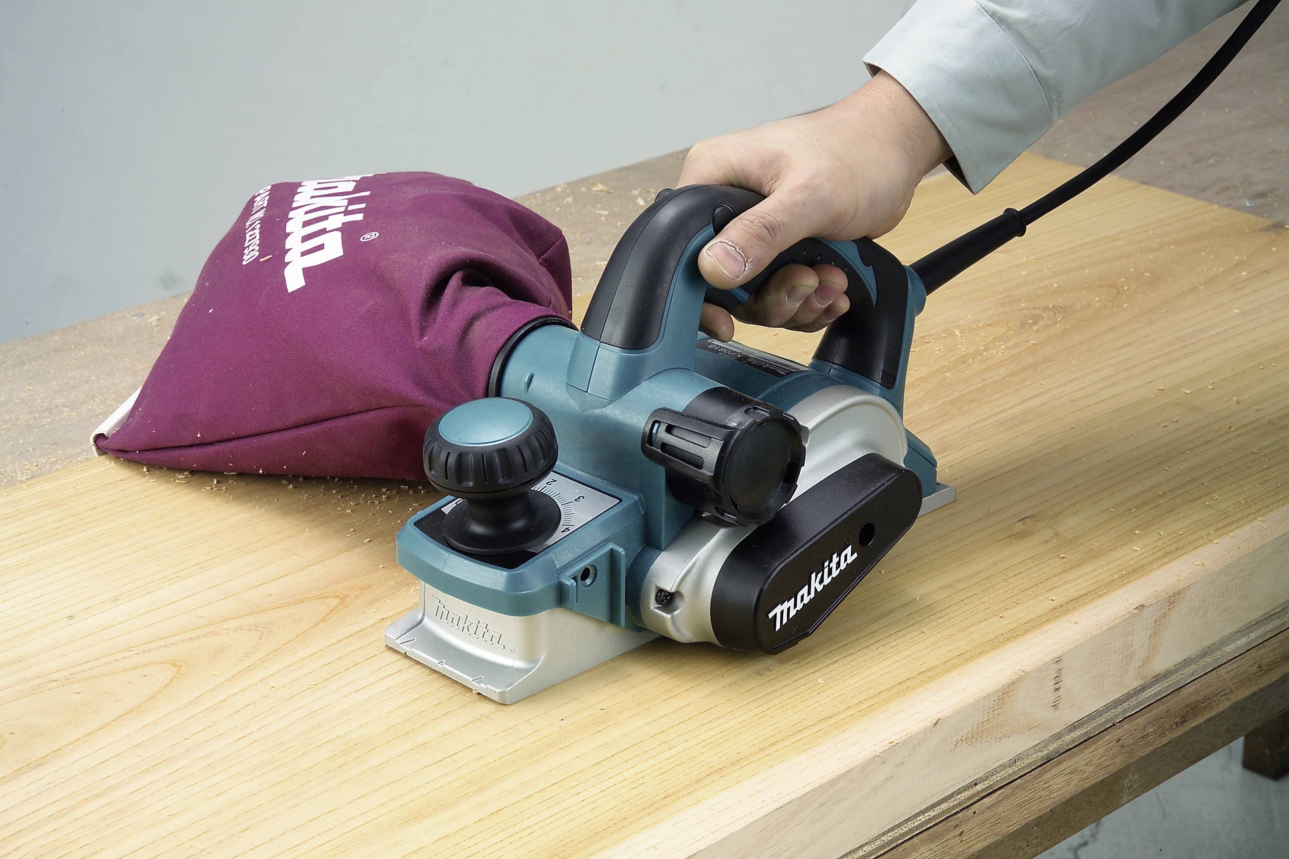 Eine Person bedient einen elektrischen Hobel, der über ein Holzbrett gleitet. Ein Staubbeutel sammelt Späne.