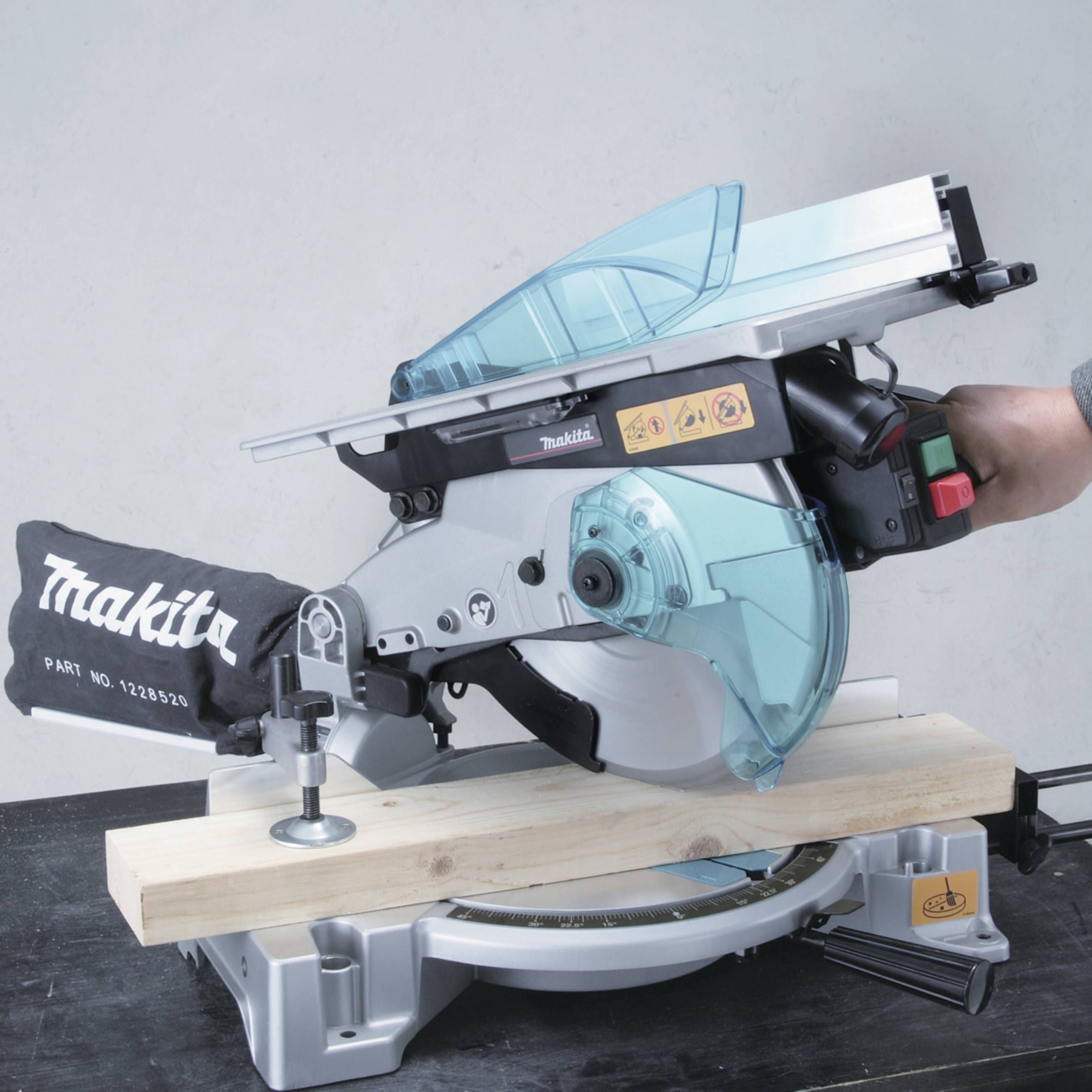 Eine Kappsäge der Marke Makita auf einem Tisch, beim Schneiden eines Holzbalkens. Eine Hand bedient die grüne Einschalttaste.