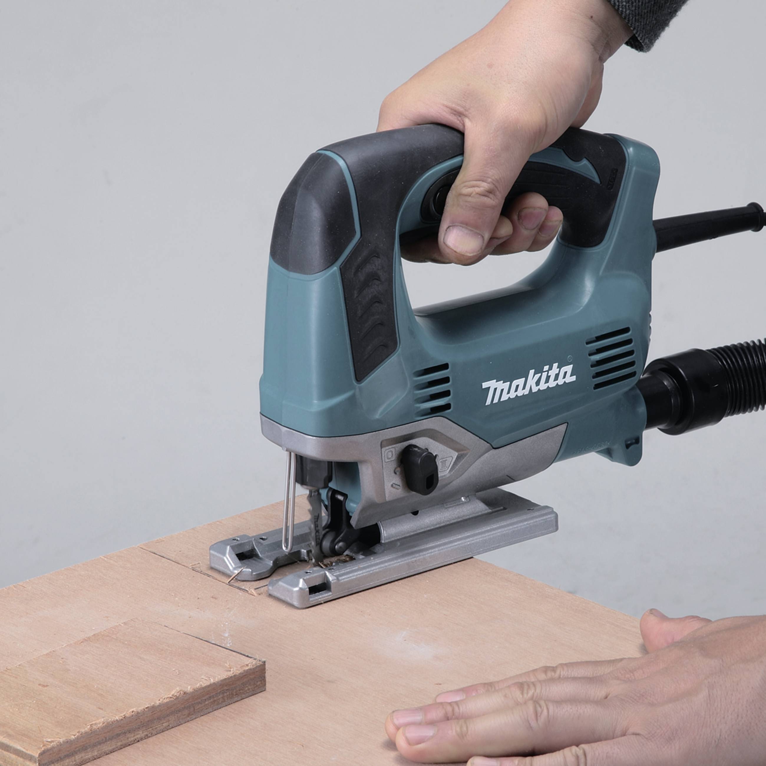 Eine Hand bedient eine elektrische Stichsäge, die ein Stück Holz auf einem Tisch schneidet.