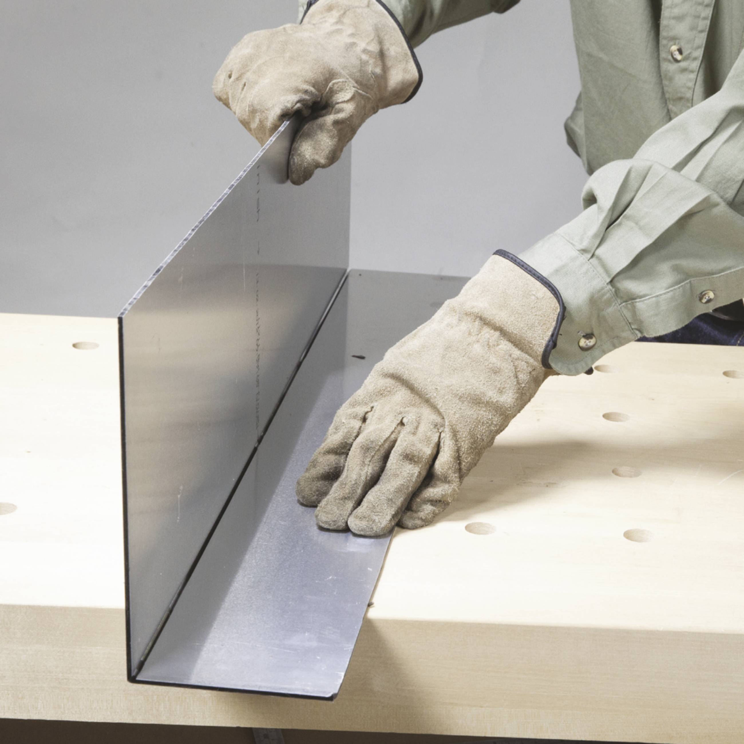 Eine Person mit Schutzhandschuhen hält ein großes Metallblech an einem Tisch. Das Blech wird gebogen oder geformt.