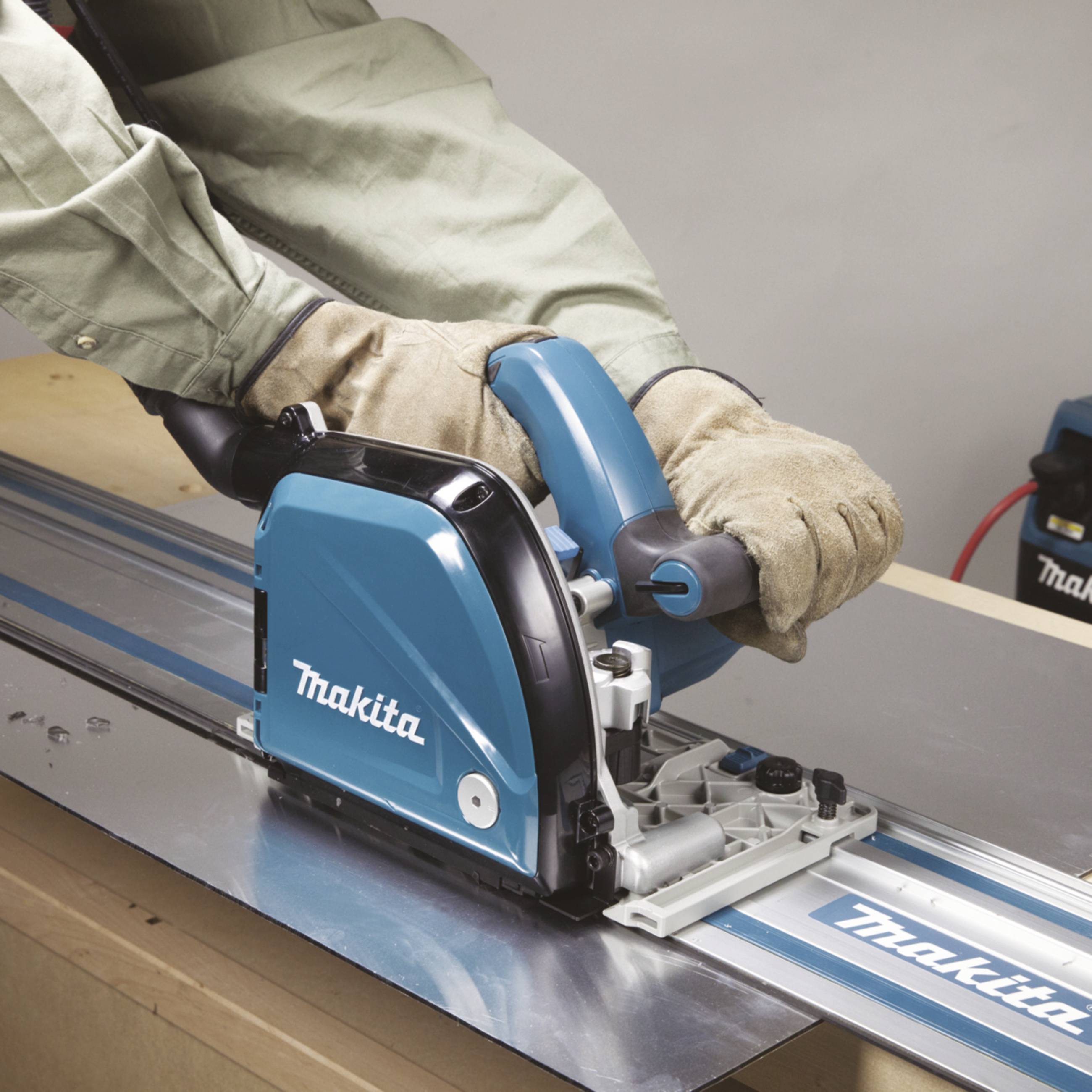 Eine Person arbeitet mit einer Tischkreissäge auf einer Holzplatte. Die Säge hat die Aufschrift 'Makita'. Die Person trägt Schutzhandschuhe.