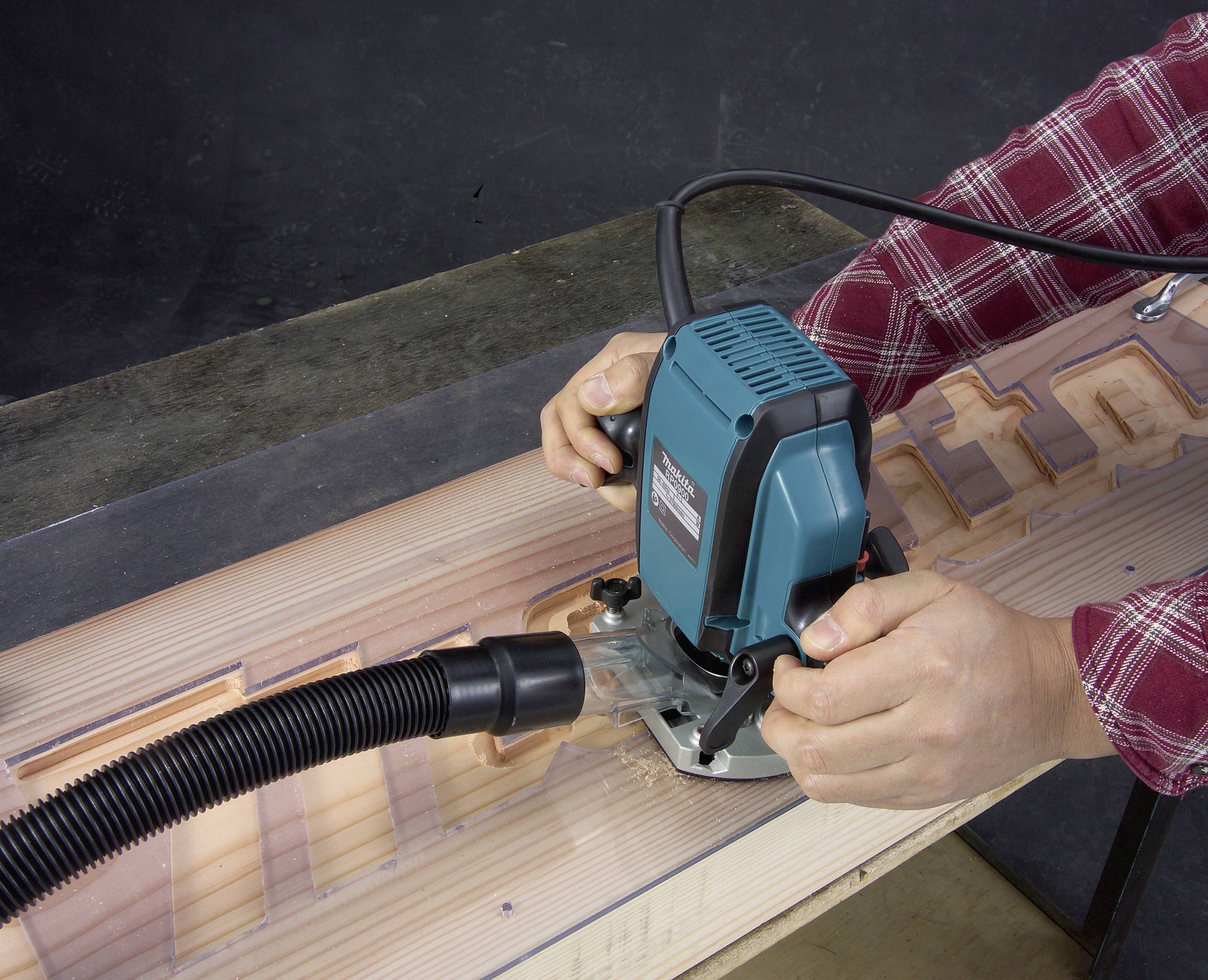 Eine Person verwendet eine Oberfräse, um Holz zu bearbeiten. Späne werden durch einen Schlauch abgesaugt. Auf dem Holz sind Buchstaben sichtbar.