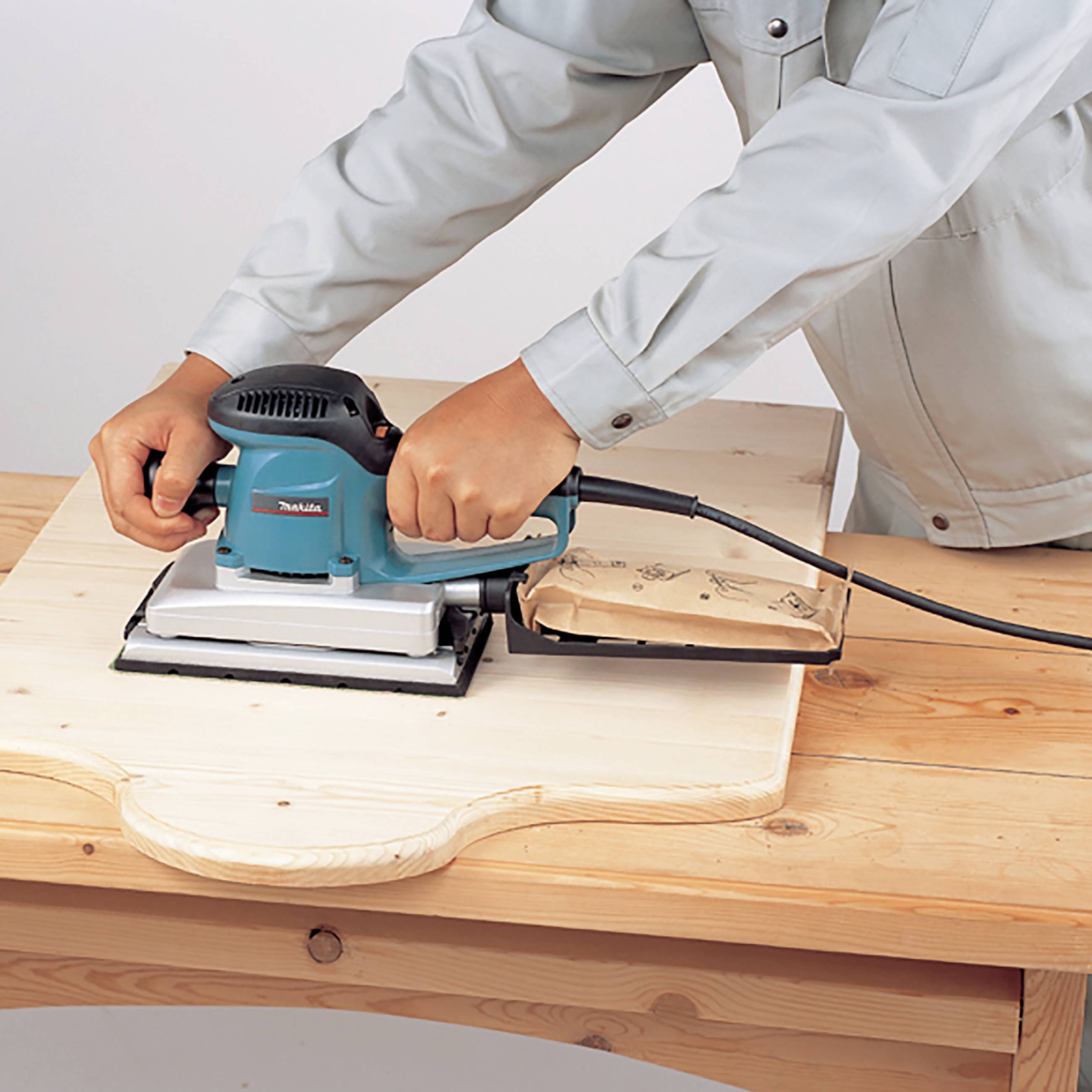 Eine Person schleift mit einem elektrischen Schleifgerät ein Holzbrett auf einem Holztisch.