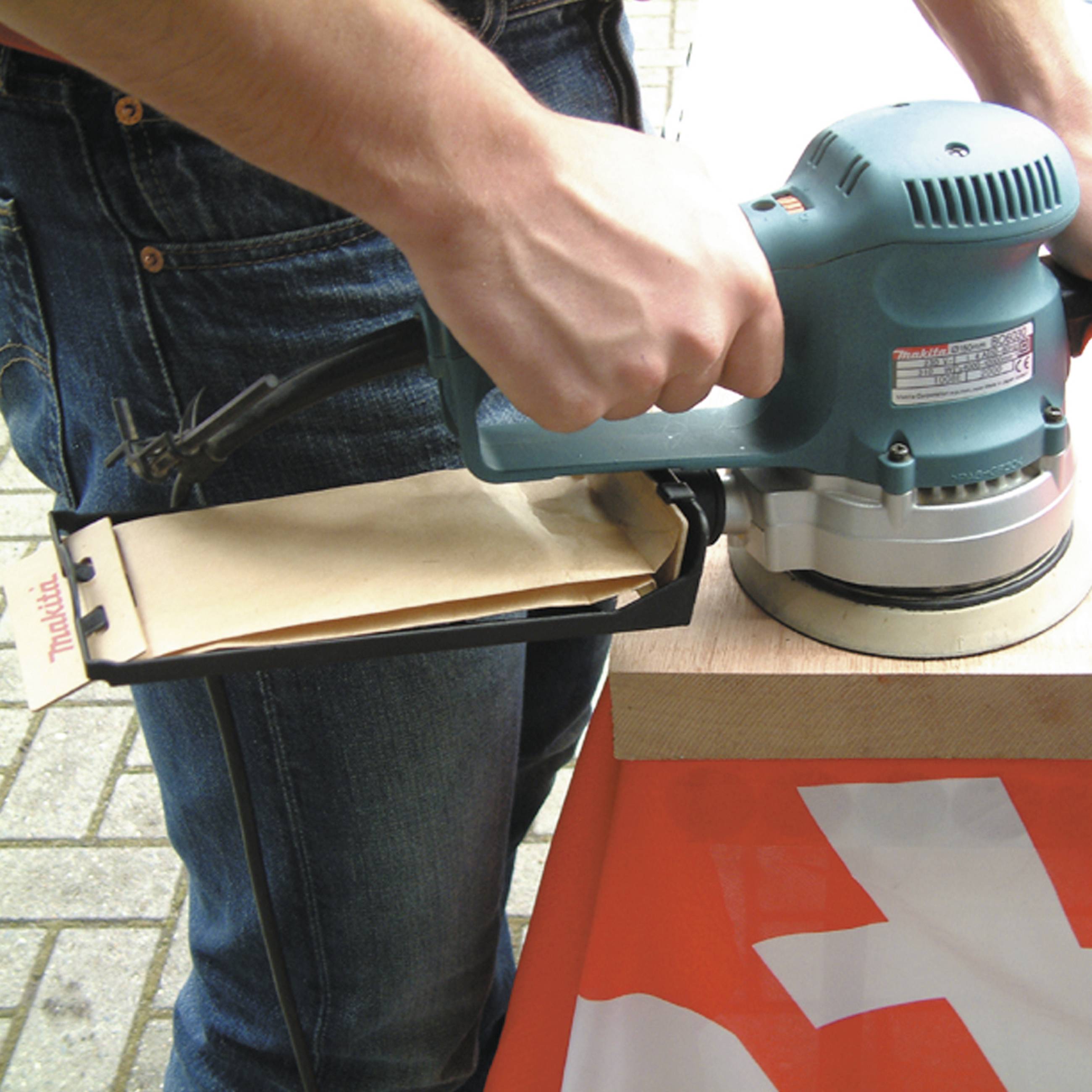 Person schleift ein Holzstück mit einer Schleifmaschine. Der Fokus liegt auf der Maschine in Aktion. Im Hintergrund sind Pflastersteine sichtbar.