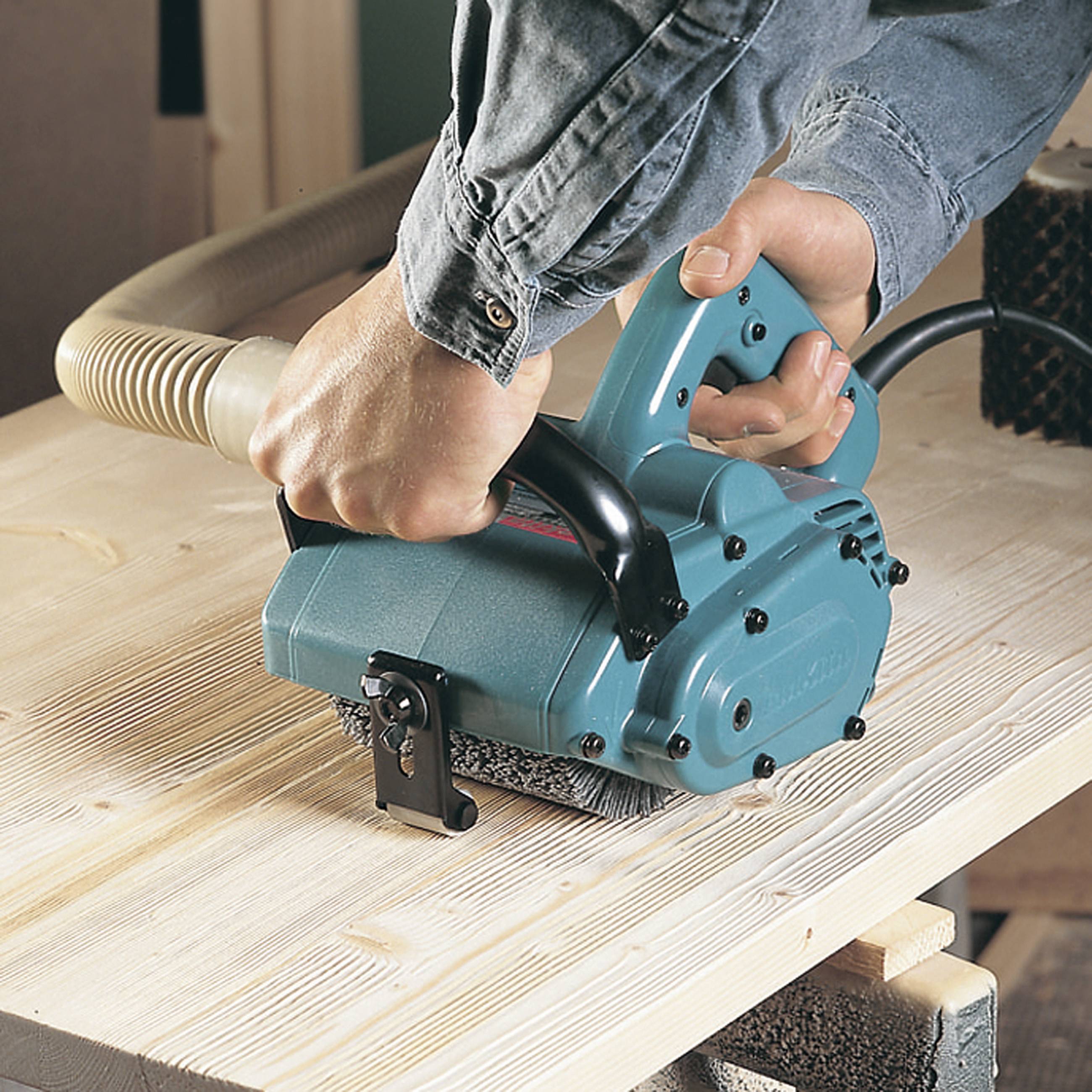 Eine Person bedient einen Elektrohobel, um eine Holzoberfläche zu glätten. Ein Absaugschlauch entfernt die Späne während des Hobelns.
