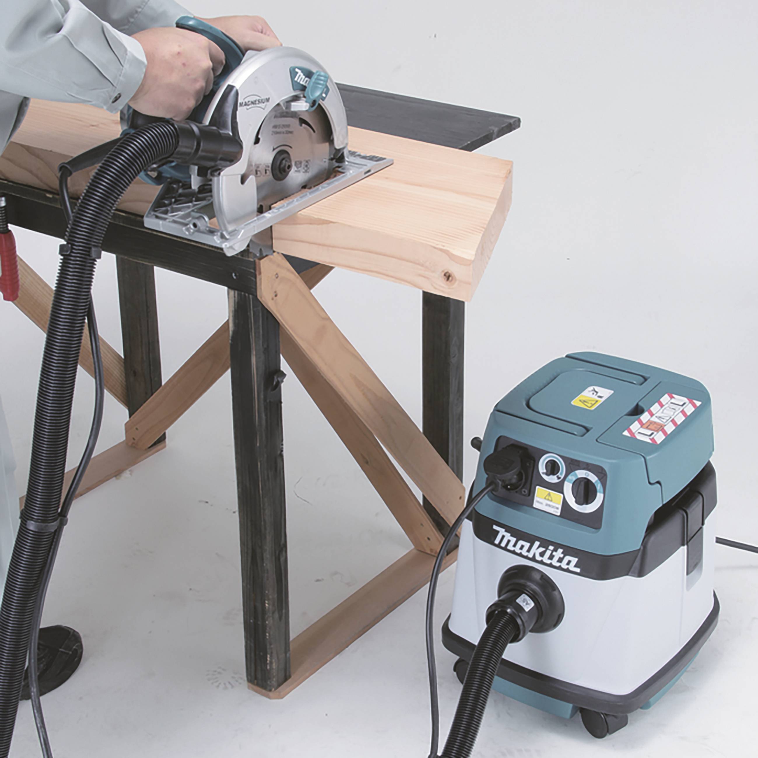 Eine Person sägt mit einer Kreissäge ein Holzbrett an einem Tisch. Ein angeschlossener Staubsauger von Makita saugt Holzspäne auf.
