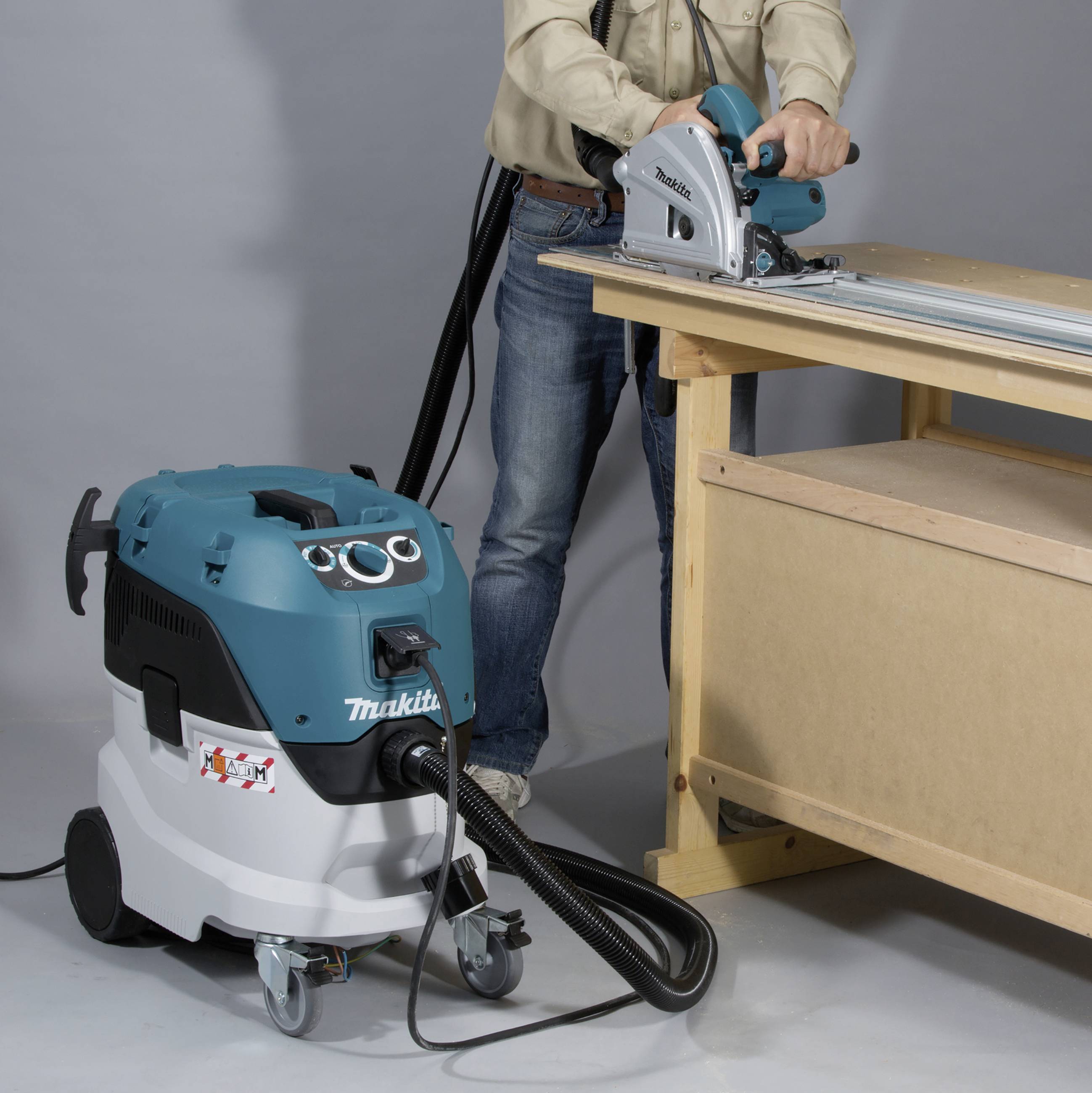 Eine Person verwendet eine elektrische Schiene und Säge auf einem Holztisch. Ein Staubsauger mit der Aufschrift 'Makita' steht daneben.