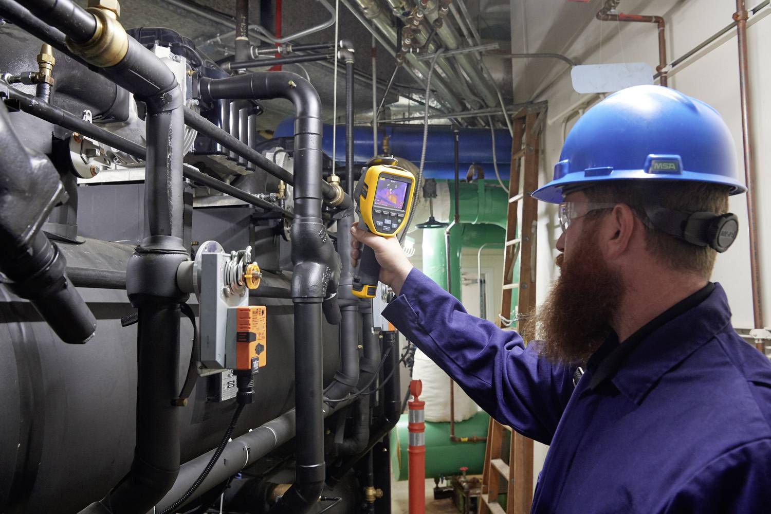 Ein Techniker in Schutzkleidung und Helm verwendet ein Wärmebildgerät, um industrielle Rohre in einem Maschinenraum zu überprüfen.