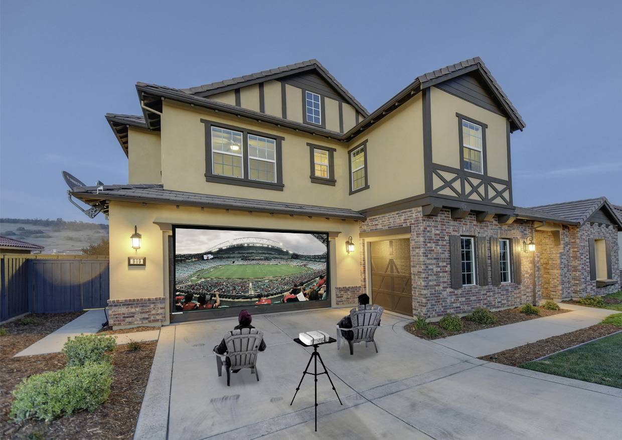 Zwei Personen sitzen auf der Auffahrt eines großen Hauses und schauen ein Fußballspiel auf einer projizierten Leinwand in der Garage.