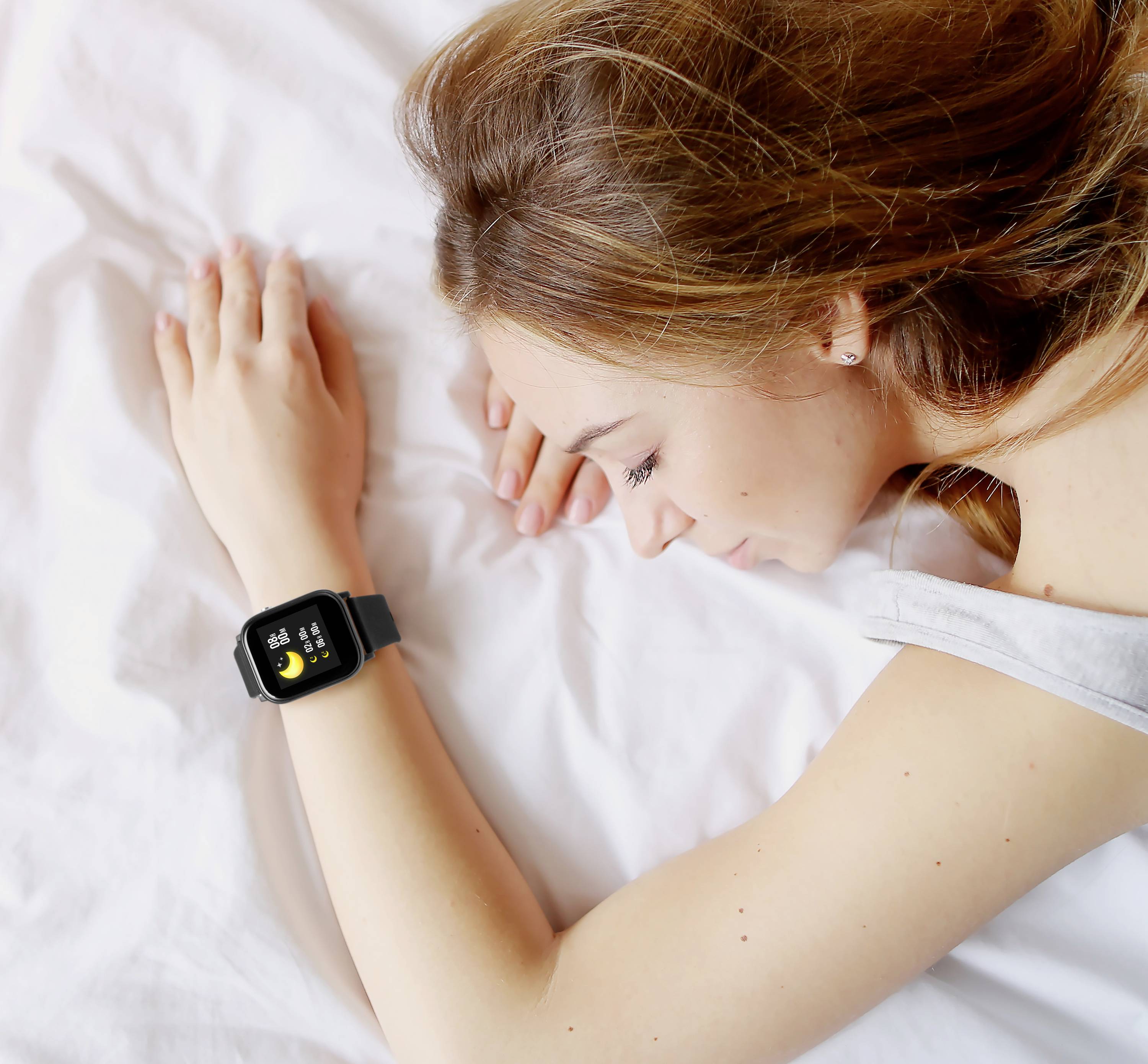 Frau liegt schlafend im Bett, trägt eine Smartwatch am Handgelenk; die Uhr zeigt Schlaftracking-Funktionen an.