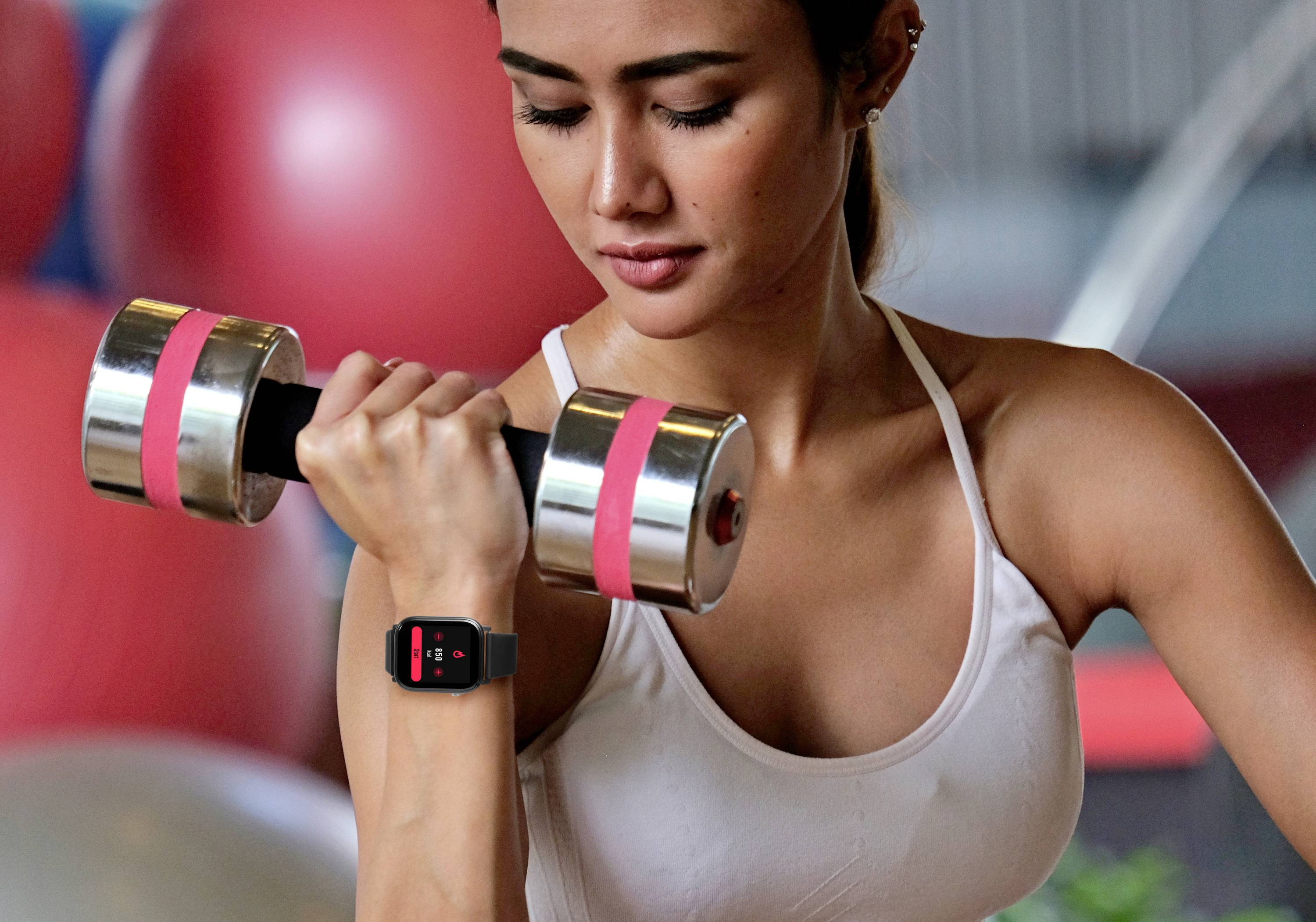 Eine Frau trainiert mit einer Hantel im Fitnessstudio. Sie trägt ein weißes Sporttop und eine Smartwatch, die Fitnessdaten anzeigt.